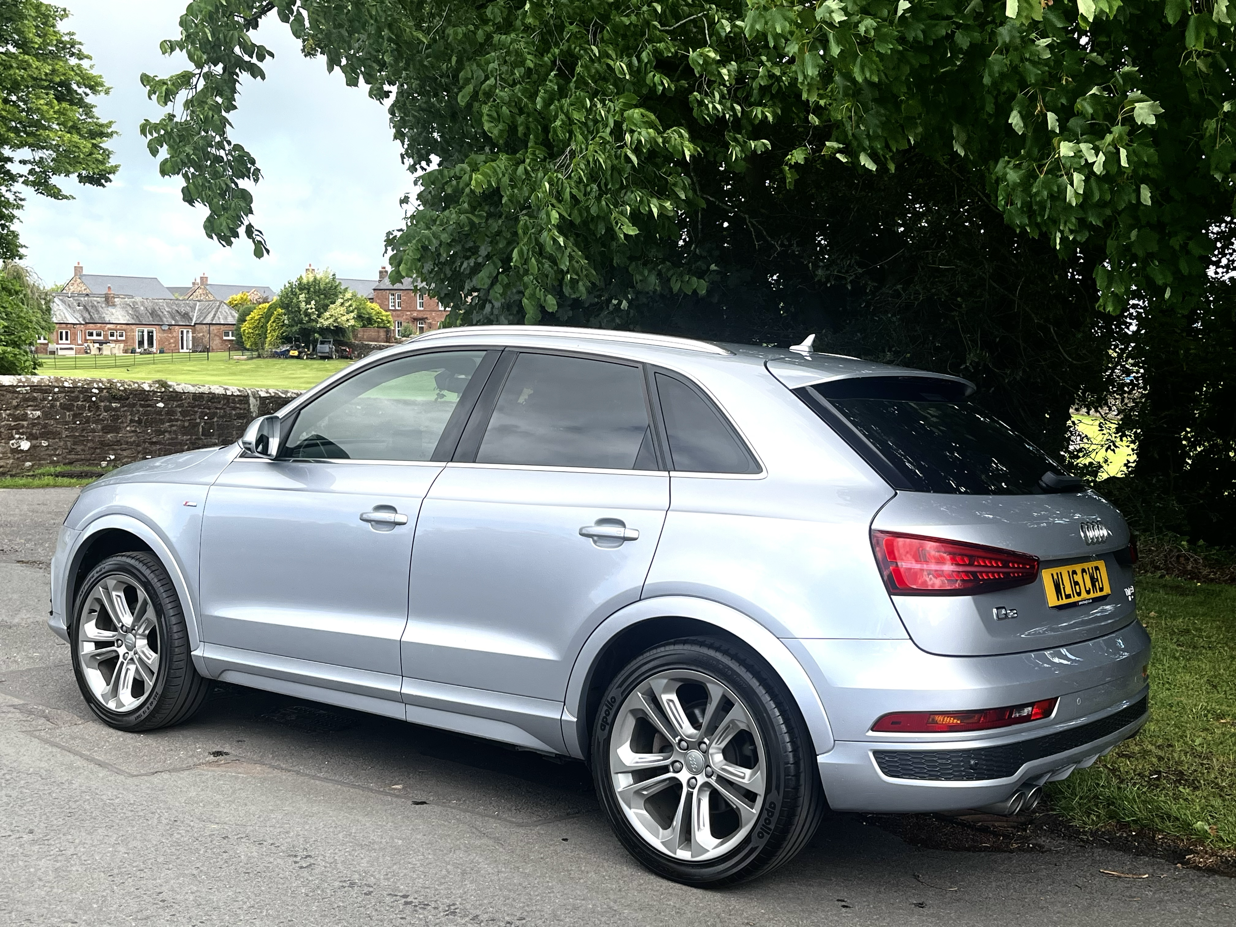 A silver Audi car parked on the side of a street next to a stone wall and green grass, with trees and houses in the background.