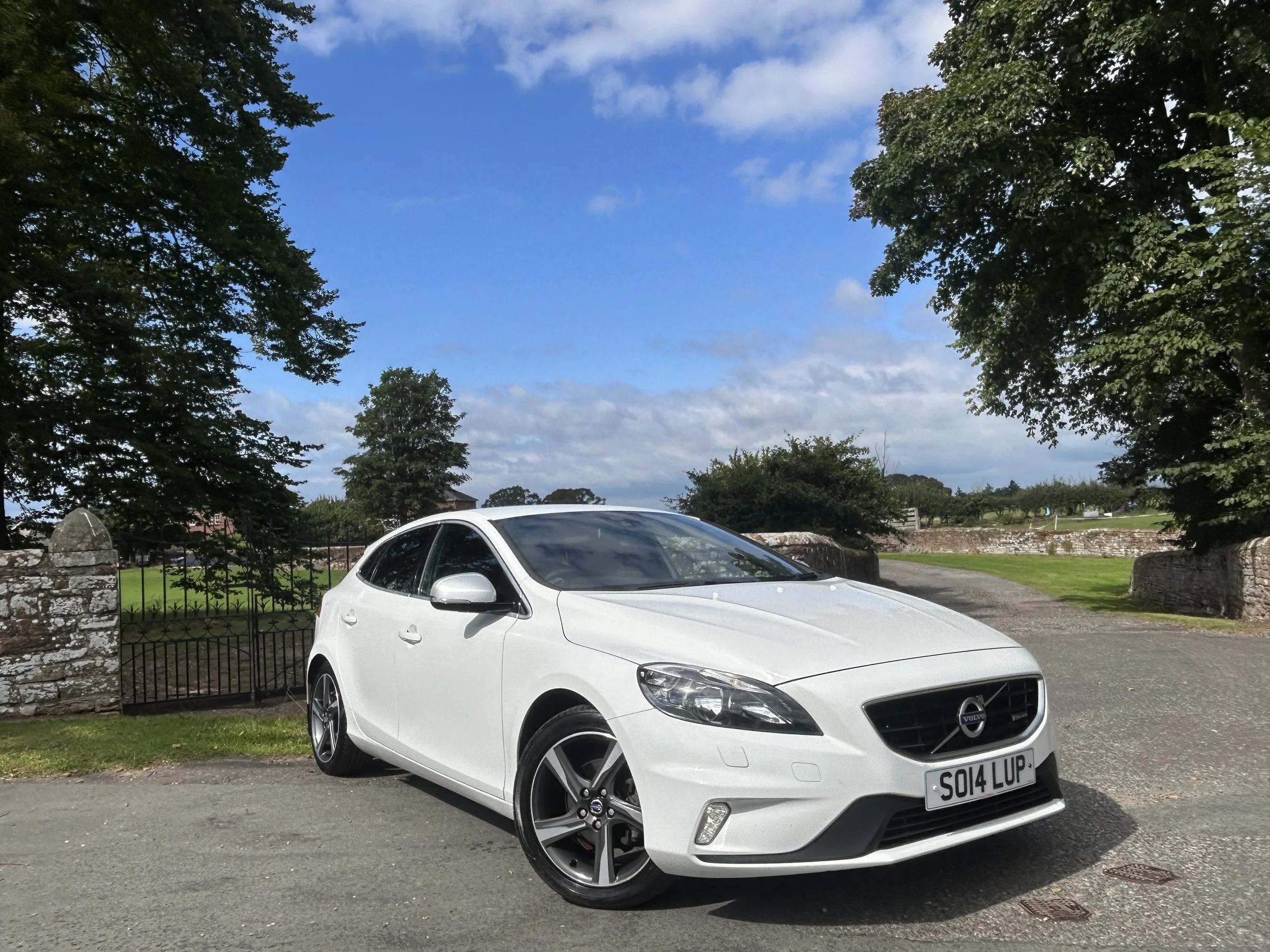 White Volvo car parked on a driveway with trees and a stone wall in the background under a partly cloudy sky.
