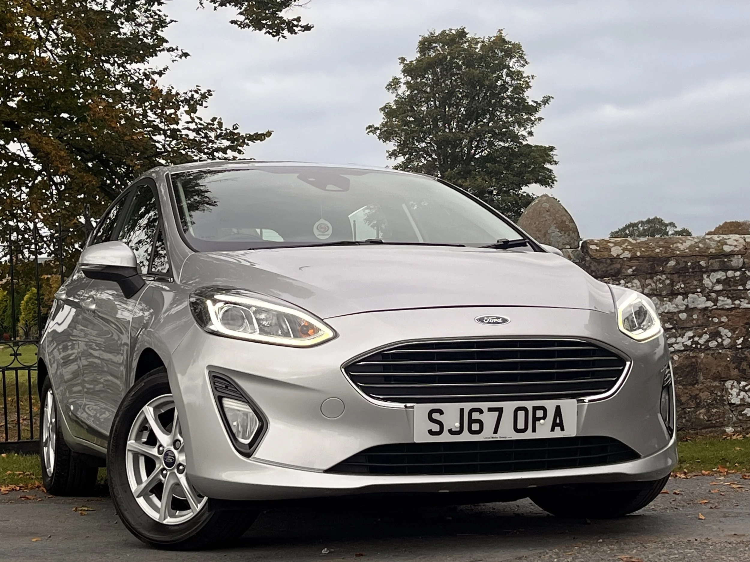 Silver Ford Fiesta hatchback parked outdoors with trees and a stone wall in the background.
