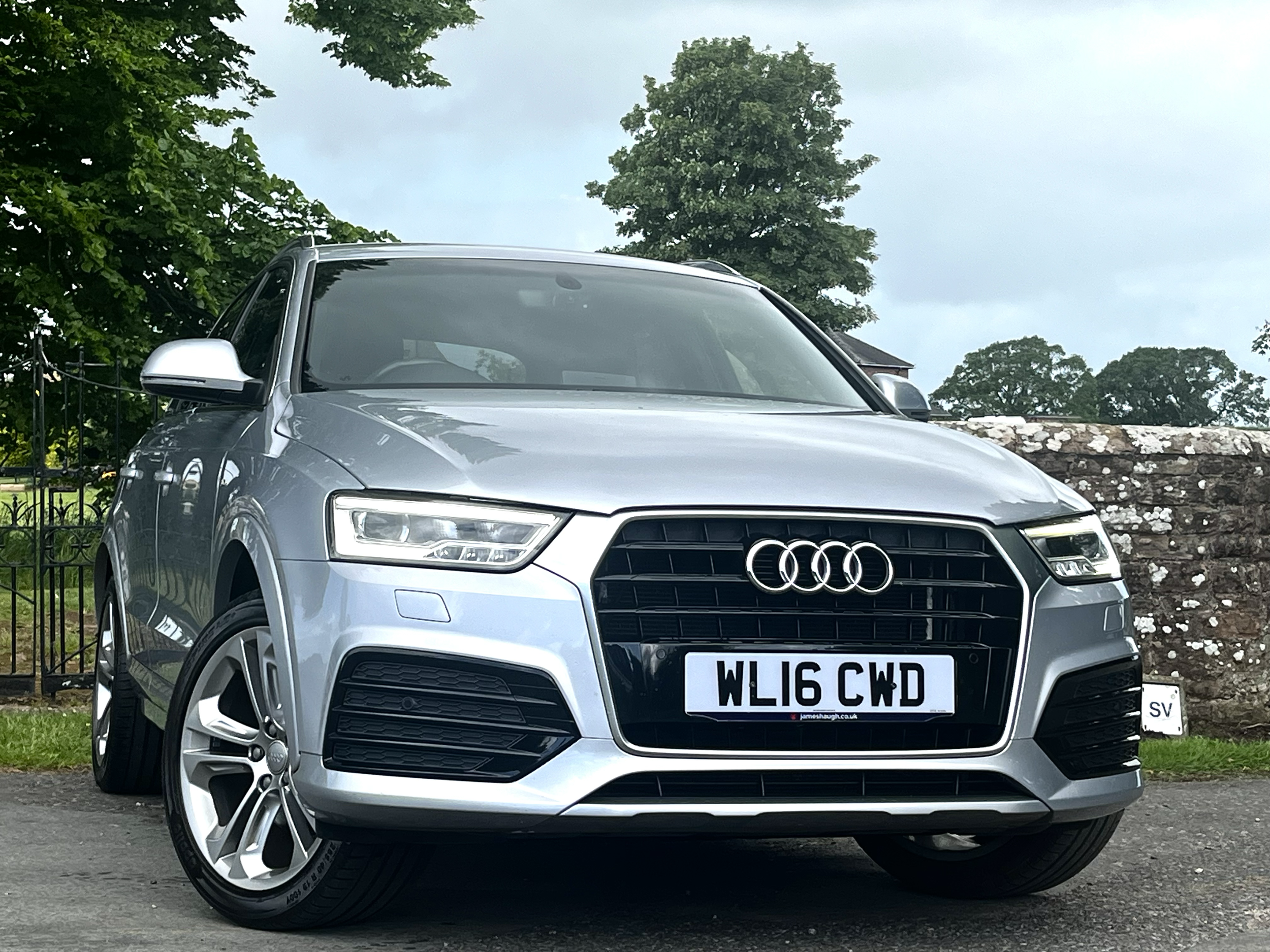 A silver Audi SUV parked outdoors with a background of trees and a stone wall. The front of the vehicle shows the Audi logo, grille, and license plate 'WL16 CWD'.