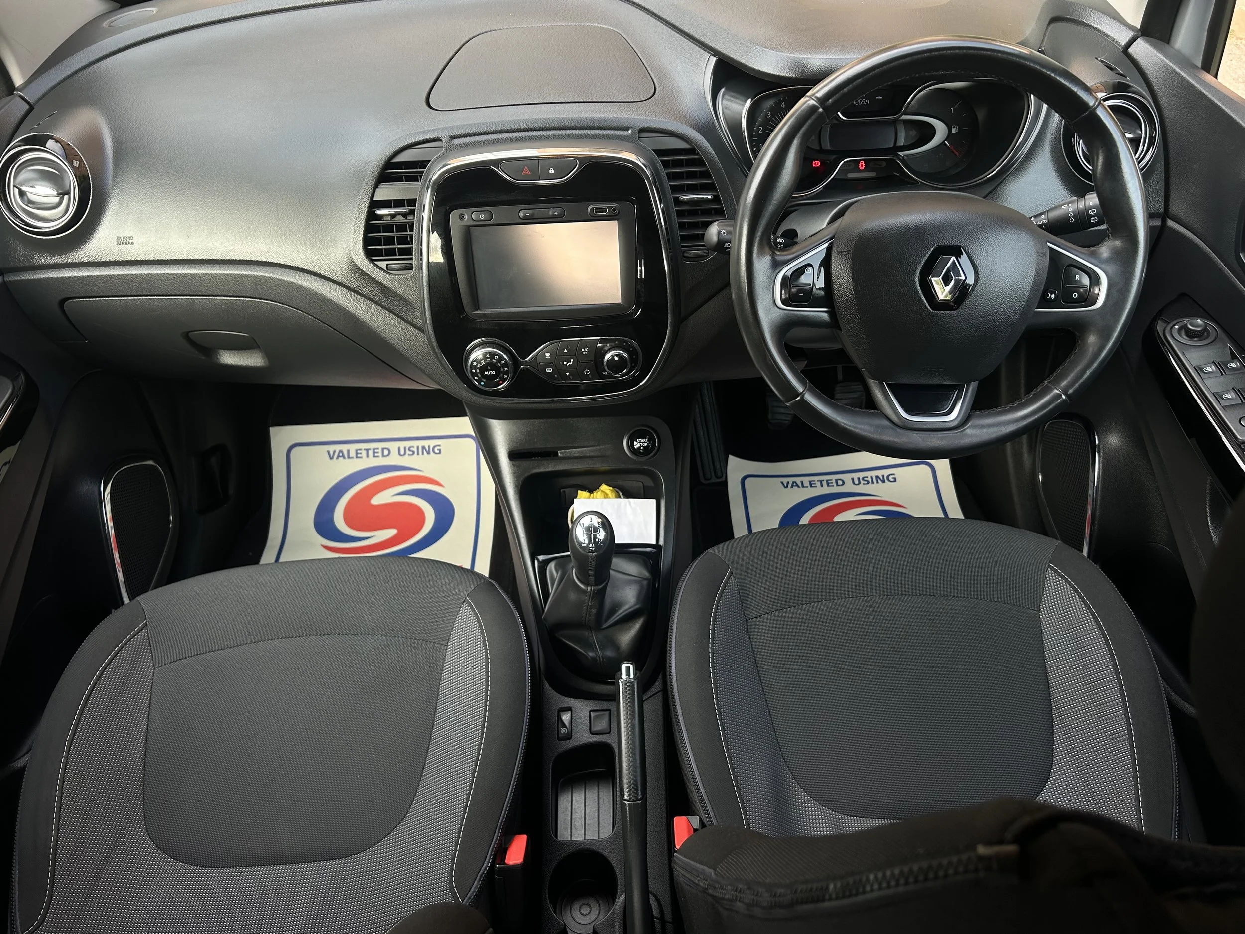 Interior of a car viewed from above, showing the dashboard, steering wheel with logo, gear shift, two front seats, and floor mats with a logo and text that says 'Valeted Using'.