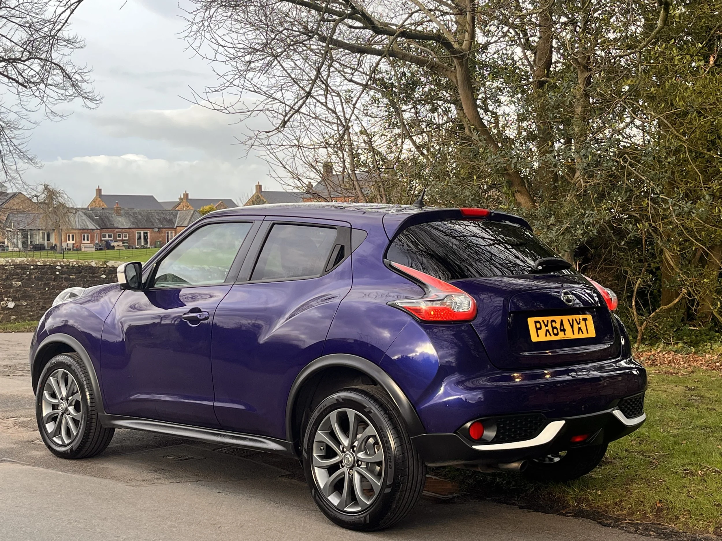 A purple compact SUV parked on a street near a grassy area with trees and houses in the background.