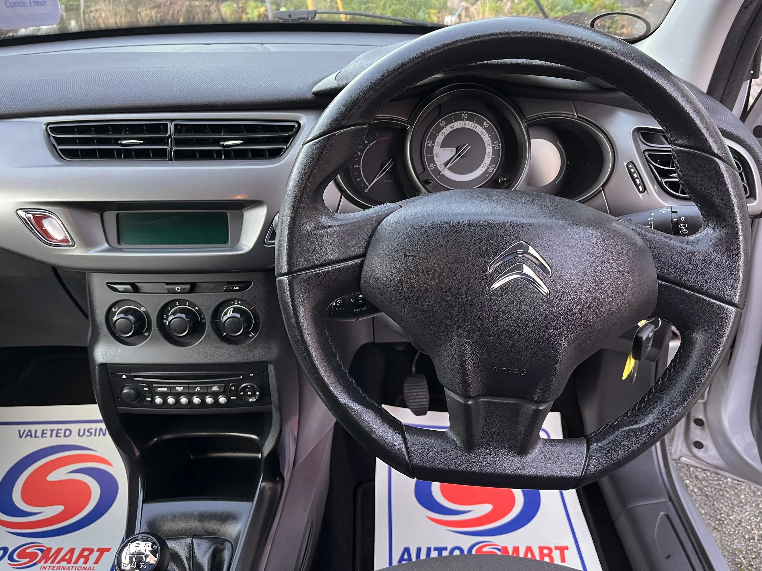Interior of a Citroën car, showing the steering wheel with the Citroën logo, the dashboard with speedometer, air vents, and control knobs, and part of the gear shift. The floor mats display the AutoSmart logo.