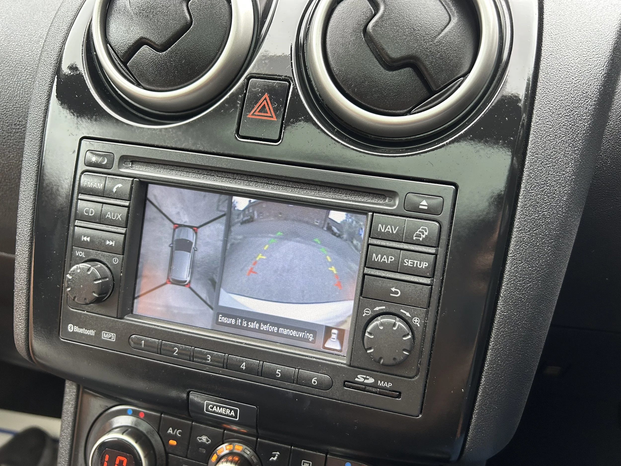 Car dashboard with a rearview camera display showing a parking guide with yellow, green, and red lines, and a top-down view of the vehicle in a parking spot.
