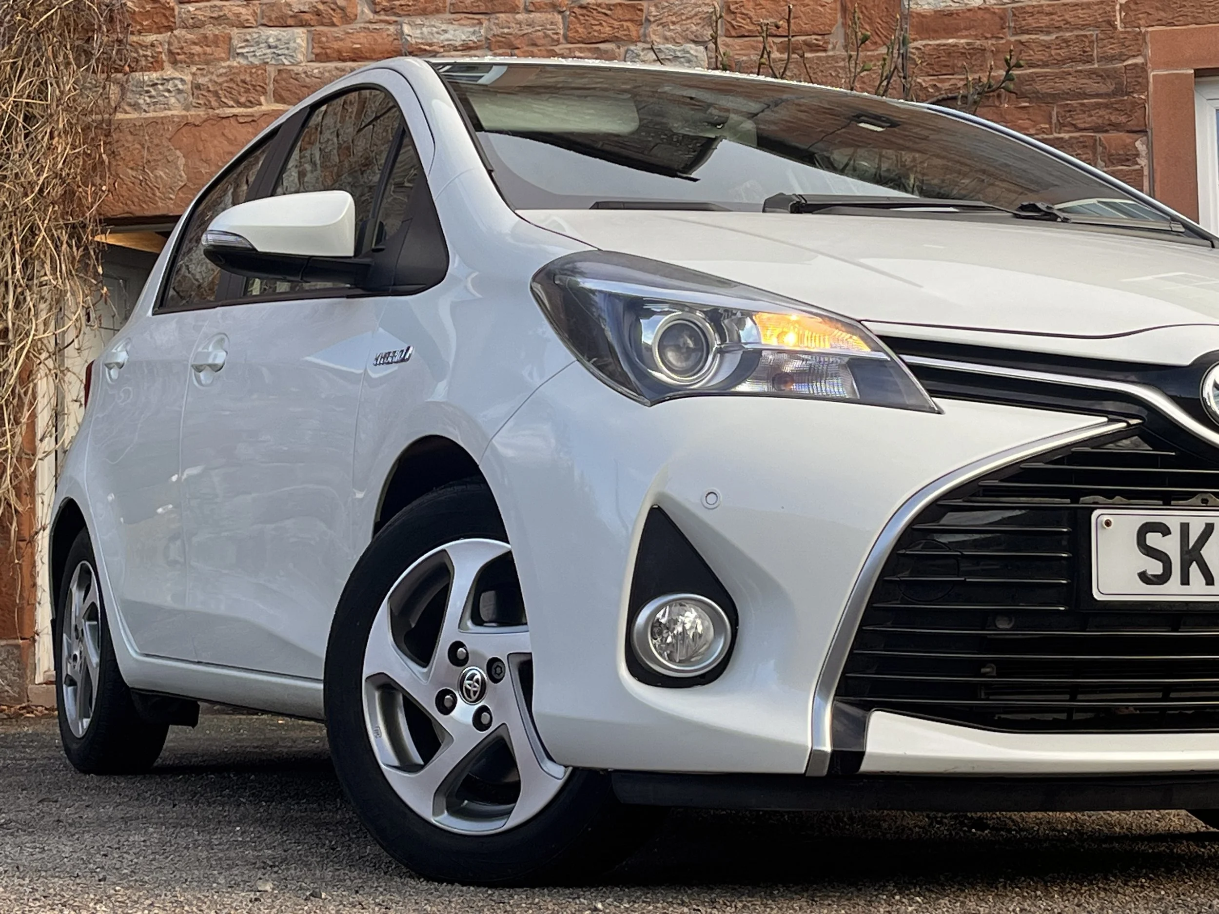 White Toyota Yaris hatchback parked on street in front of a brick wall with plants