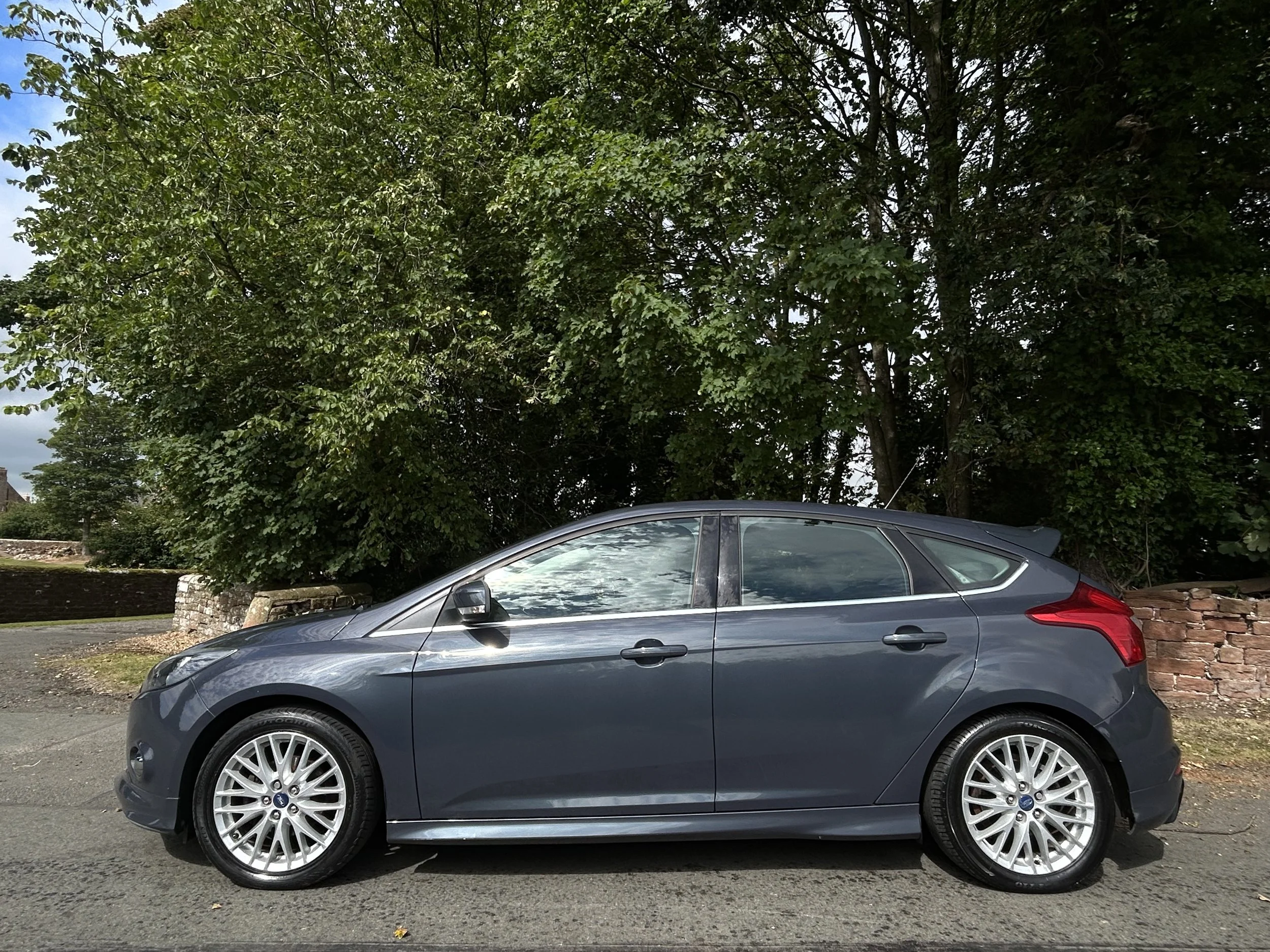 Gray hatchback car parked on the side of the road with green trees and a brick wall in the background.