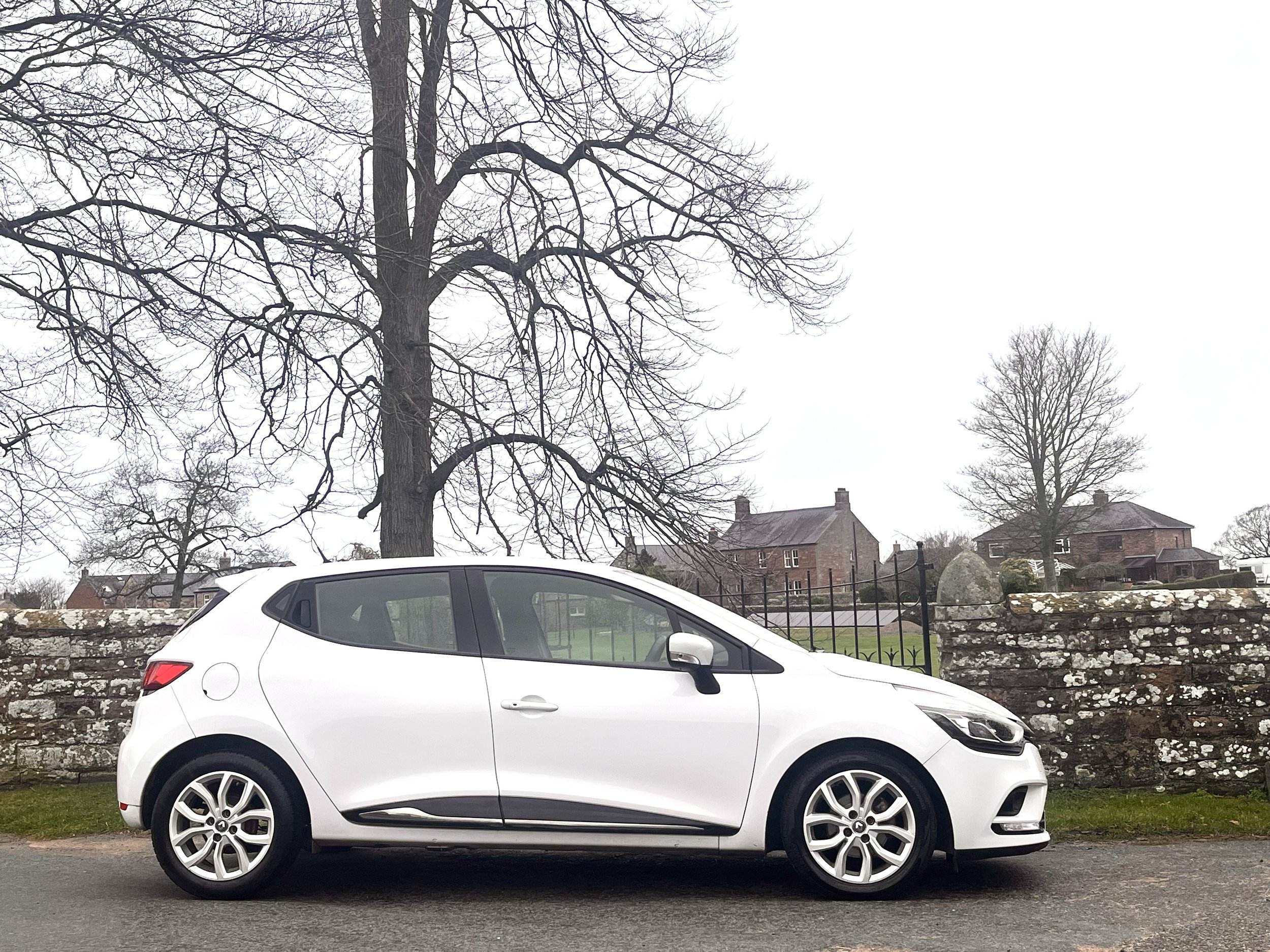 A white hatchback car parked on the side of a street in front of a stone wall, with leafless trees and houses in the background on a cloudy day.