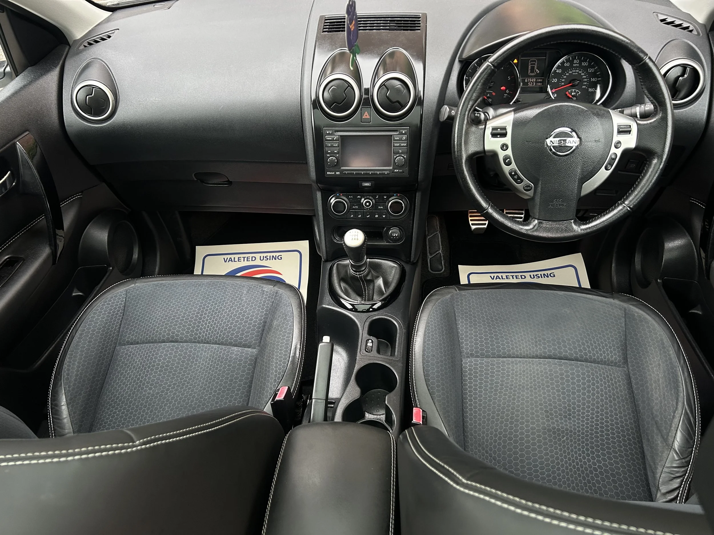 Interior view of a Nissan vehicle showing the dashboard, steering wheel, gear shift, and two front seats with black upholstery and white stitching.