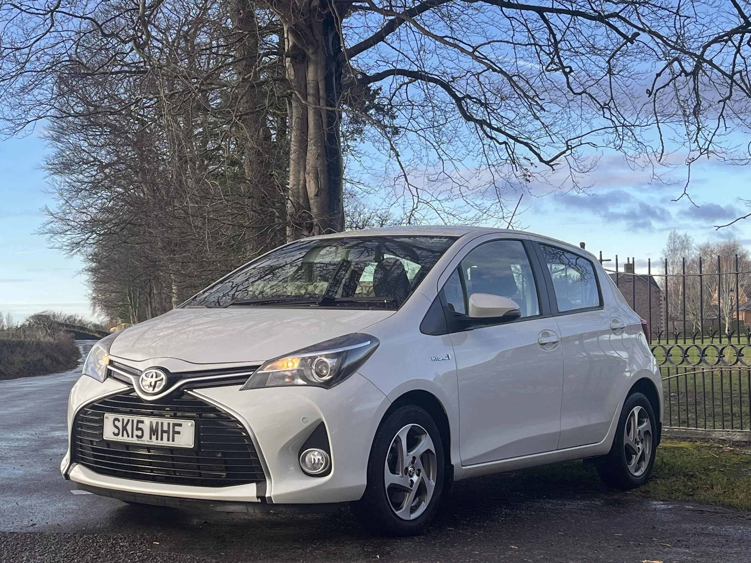 Silver Toyota Aygo parked on a wet pavement with trees and a fence in the background on a partly cloudy day.