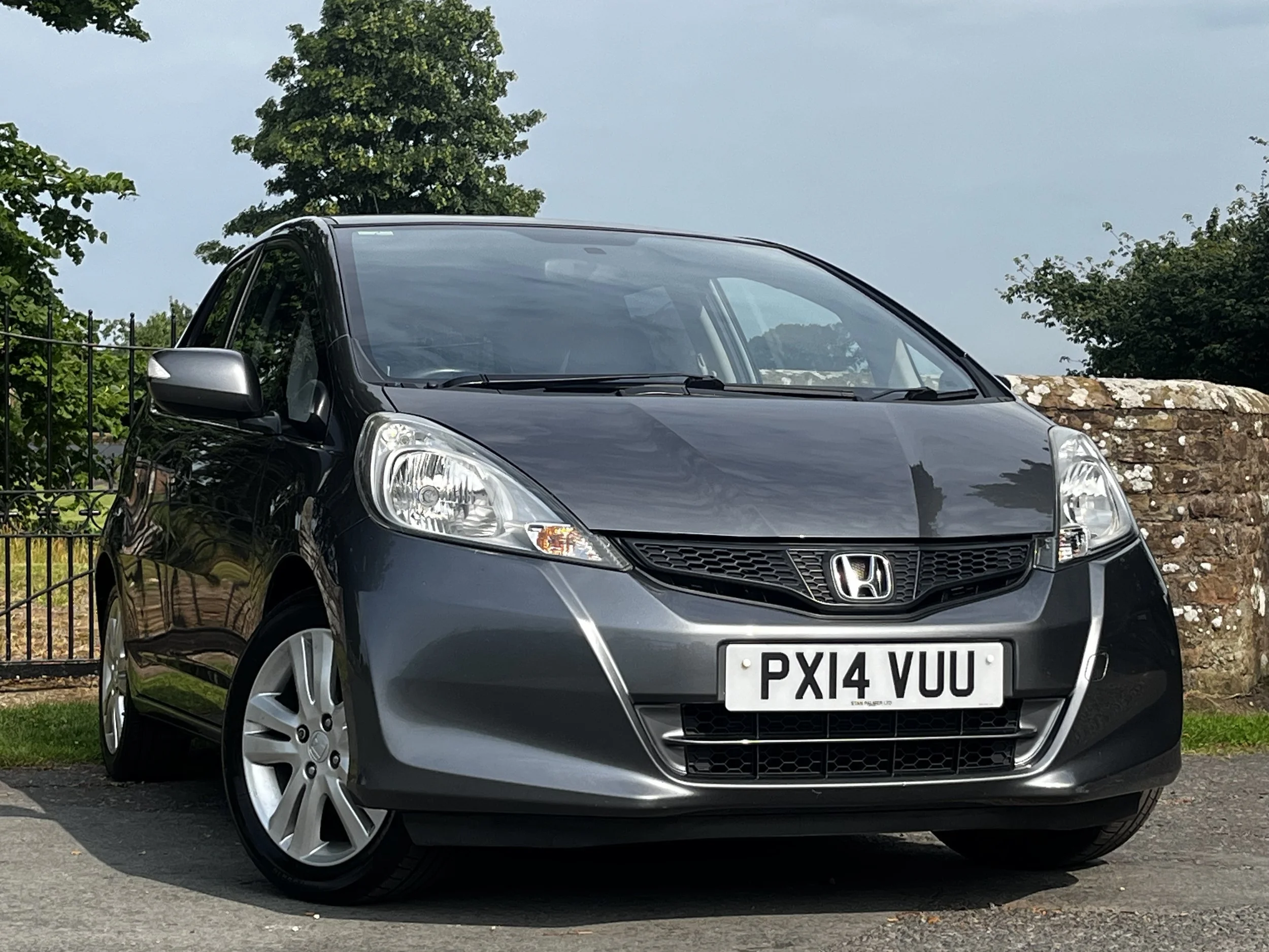 A black Honda Jazz car parked outdoors with trees and a stone wall in the background. The car has a UK license plate reading PX14 VUU.