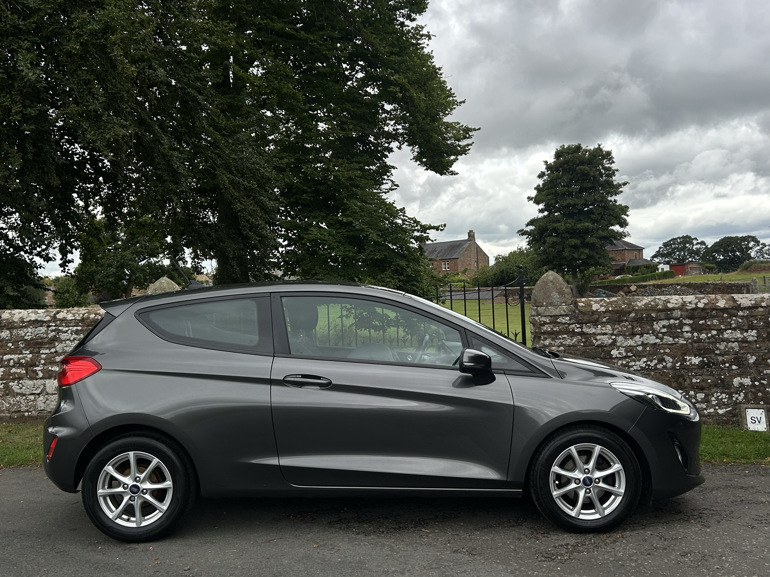 A silver Ford Fiesta parked on a gray street in front of a brick wall with large trees in the background, under a cloudy sky.