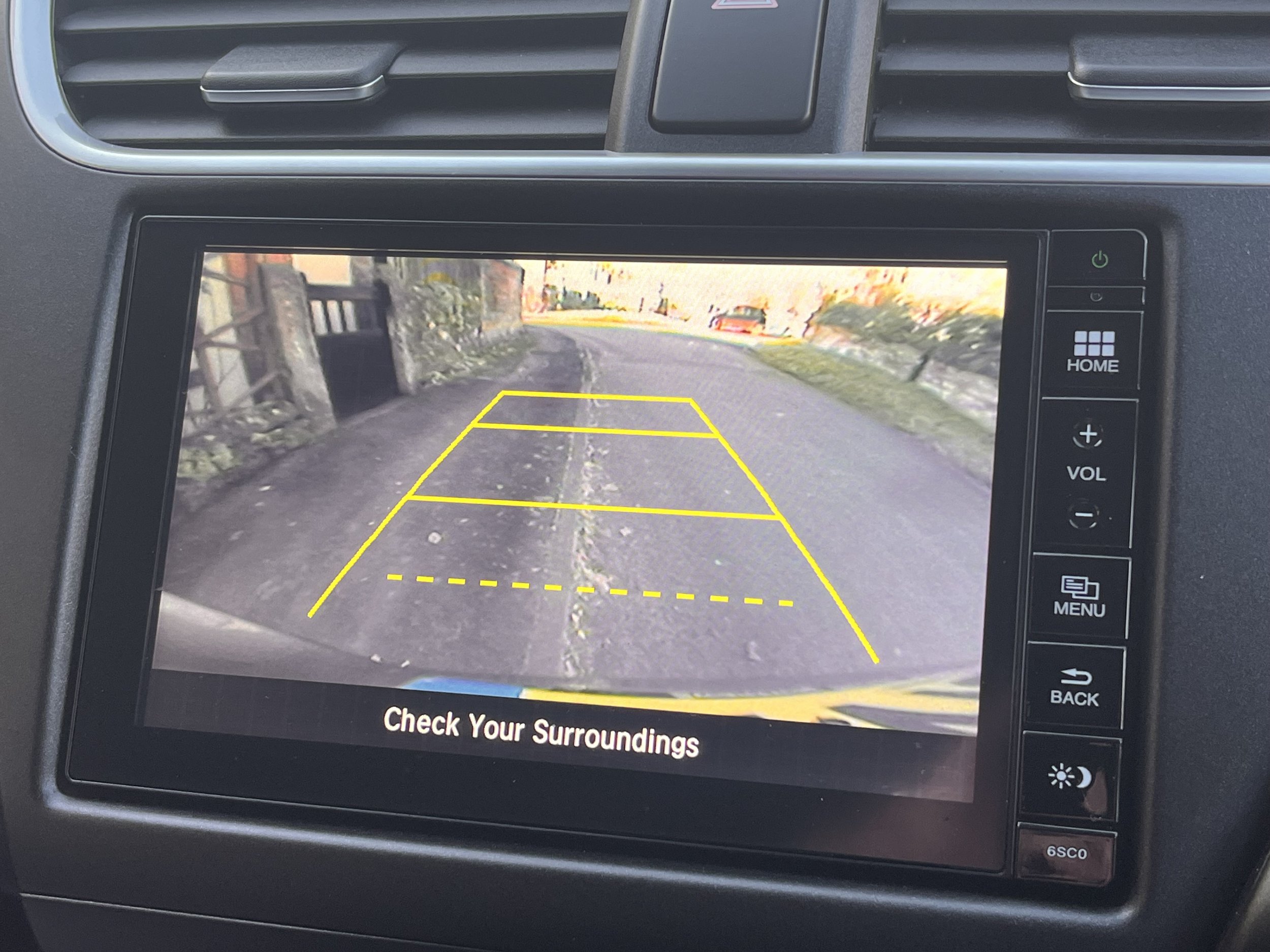 Car's rearview camera display showing a driveway with yellow parking guide lines and a caution message to check surroundings.