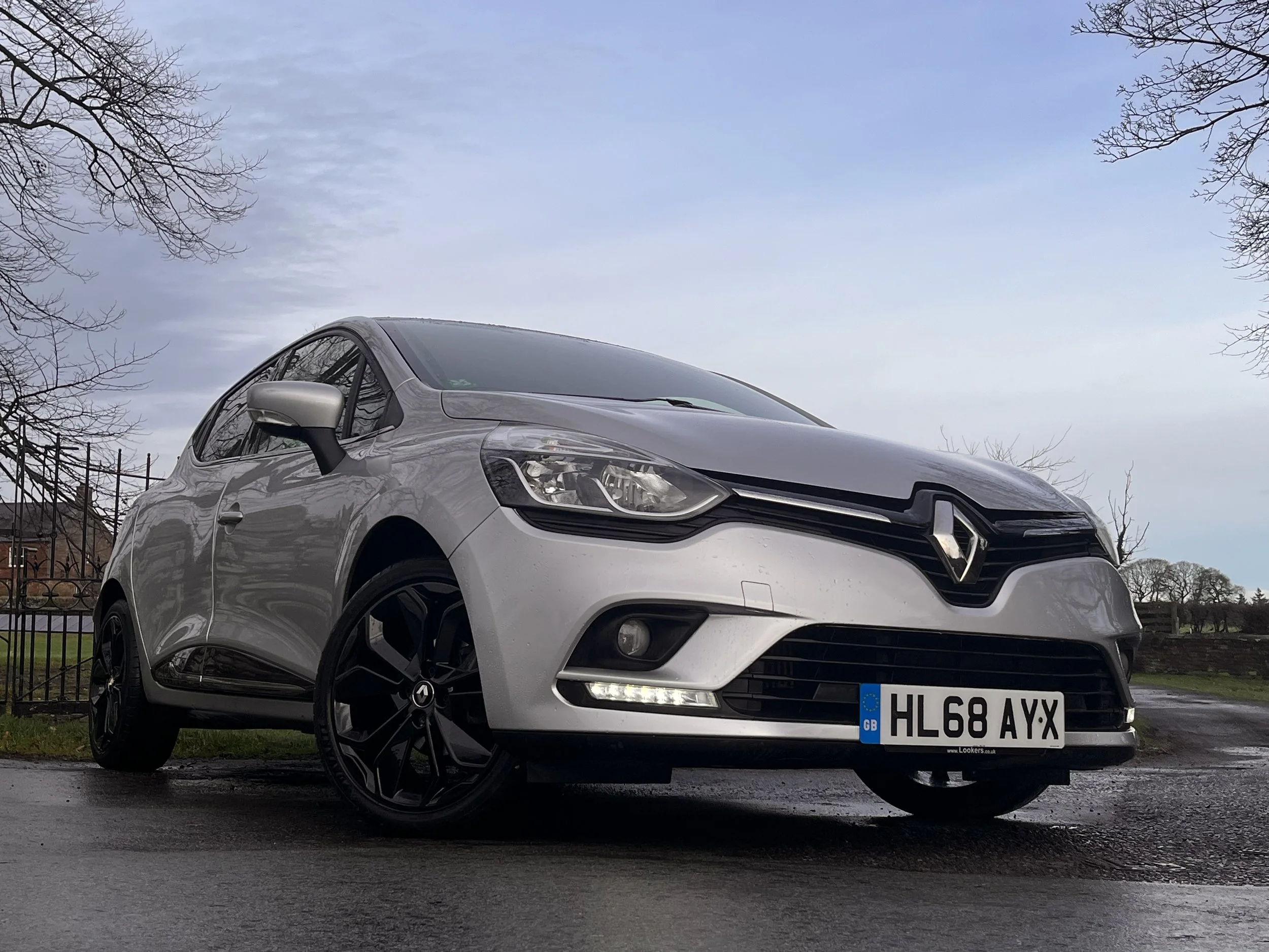Silver Renault hatchback parked on a wet road with a cloudy sky and leafless trees in the background.