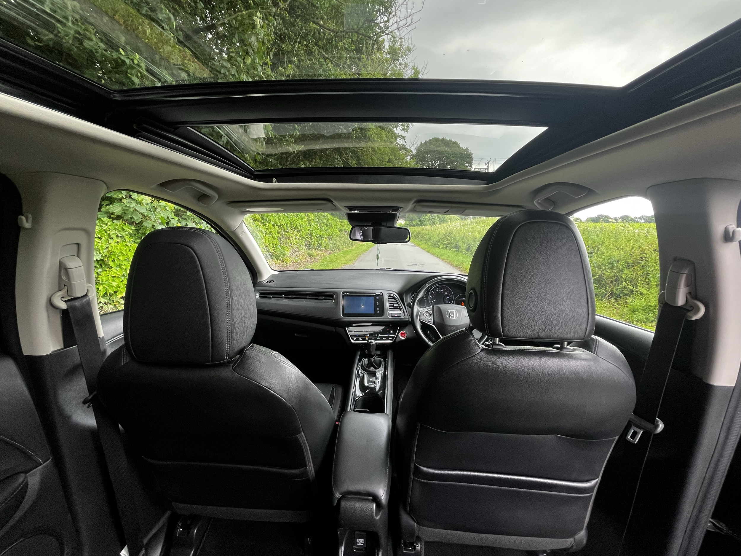 View from inside a vehicle with a sunroof, showing the dashboard, steering wheel, front seats, and a road with green trees and cloudy sky outside.