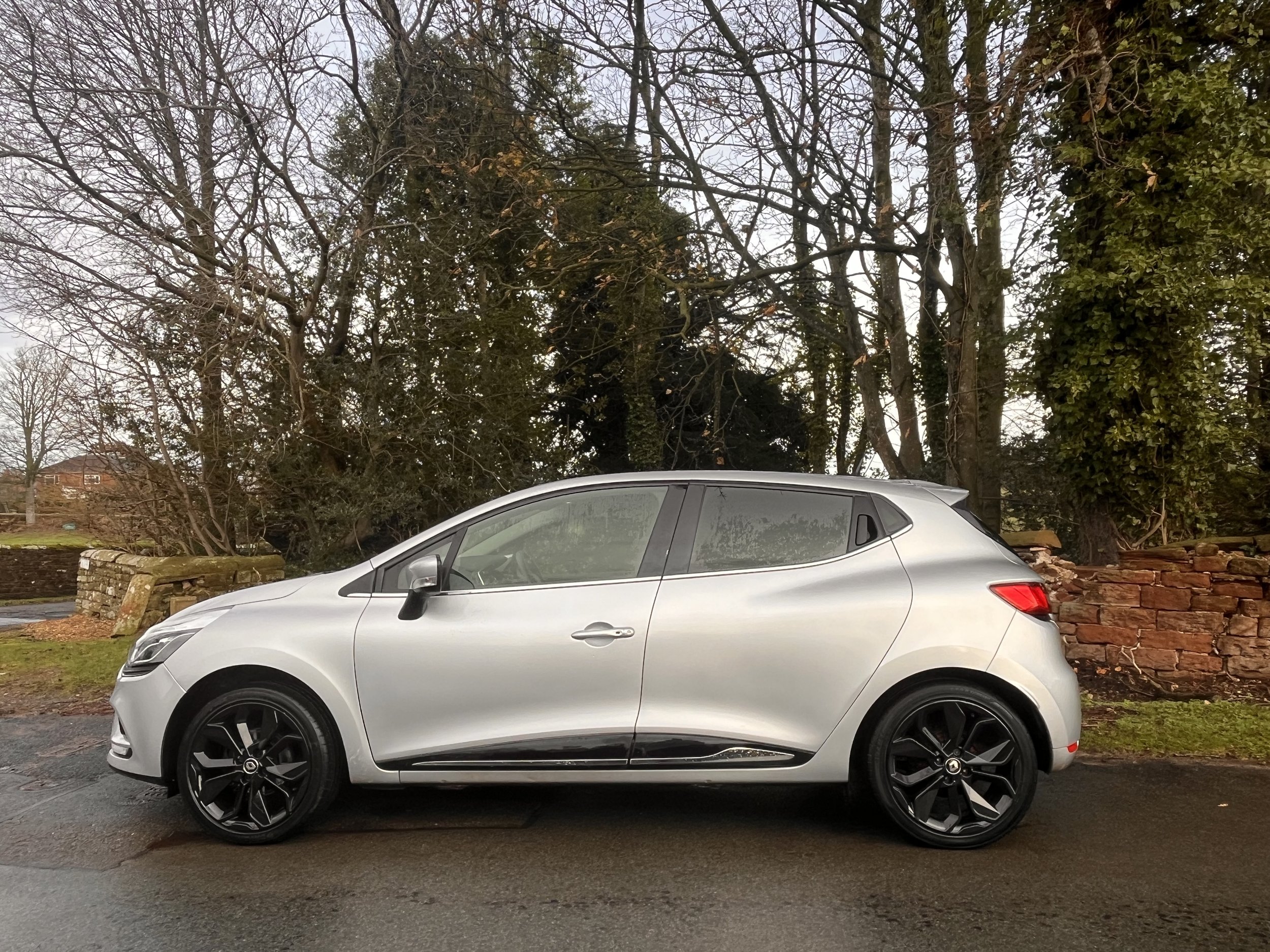 A silver hatchback car parked on a wet road with trees and a brick wall in the background.