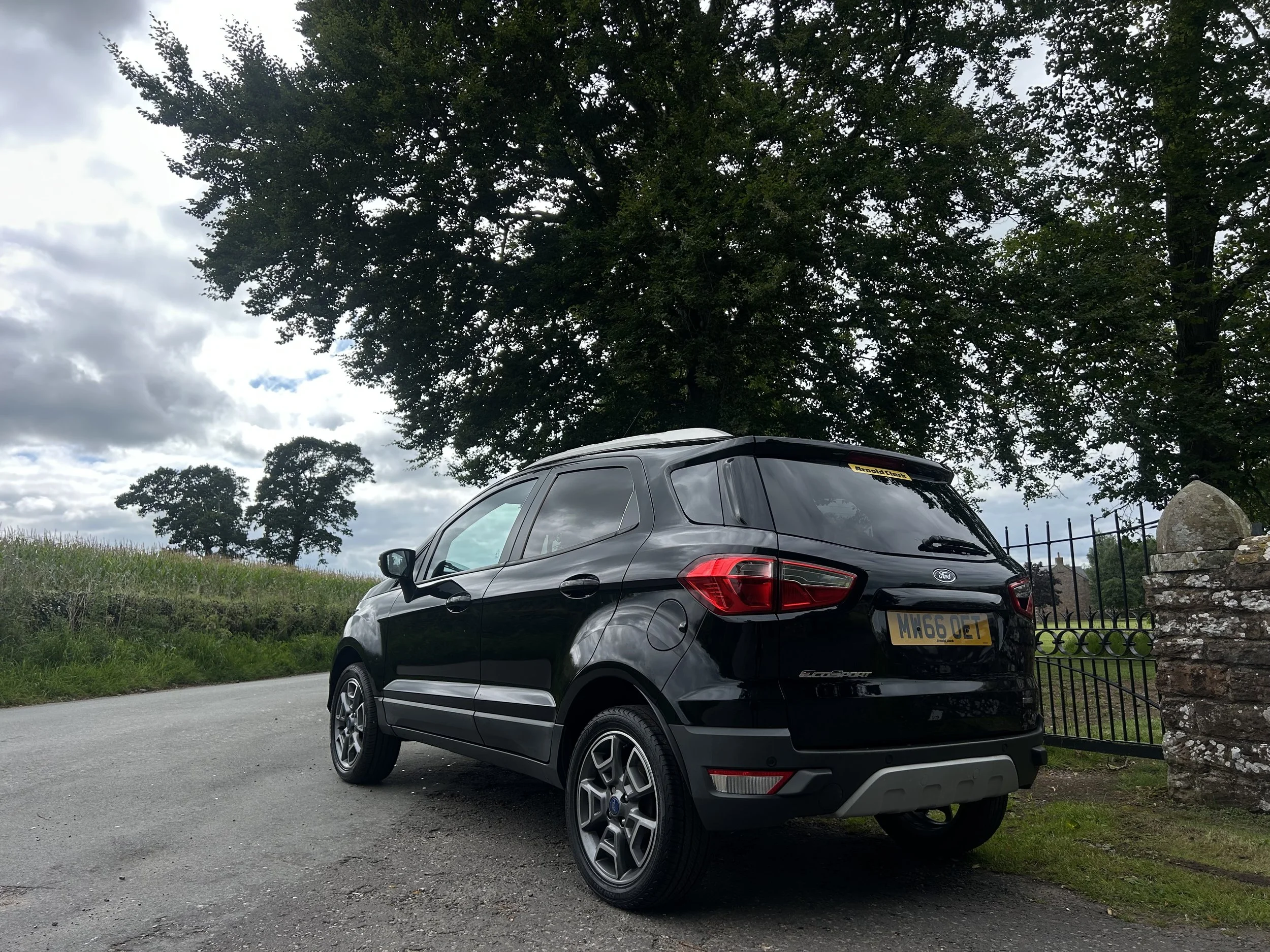 Black Ford EcoSport parked on a gravel driveway next to a stone and iron gate, with trees and a cloudy sky in the background.