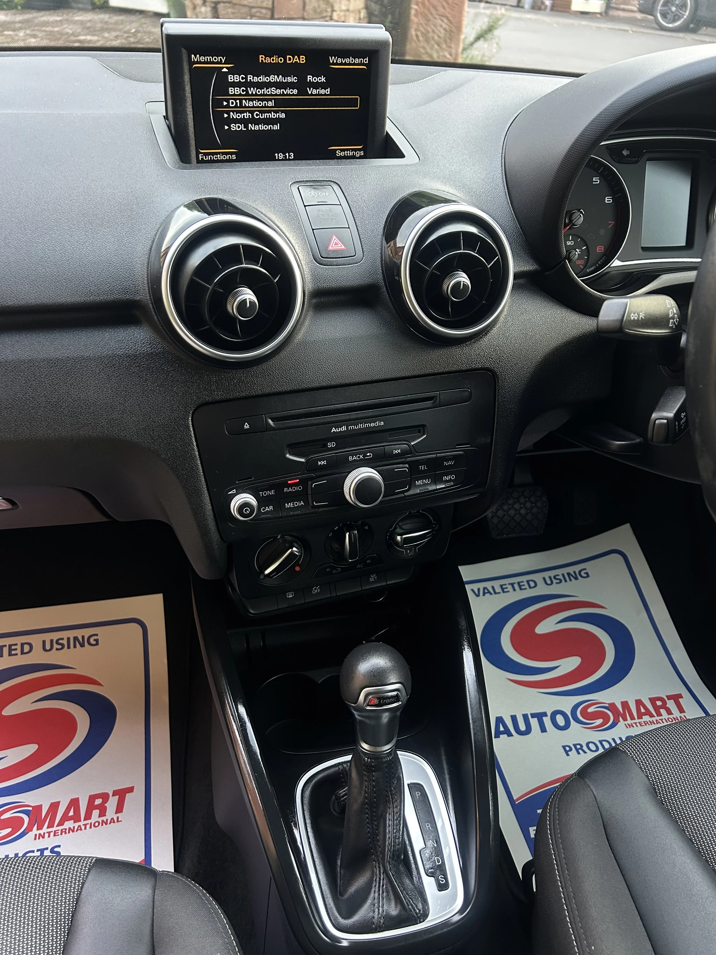 The interior of a car showing a dashboard with a central control panel, an automatic gear shift, two round air vents, a multimedia display, and floor mats branded with AutoSmart.