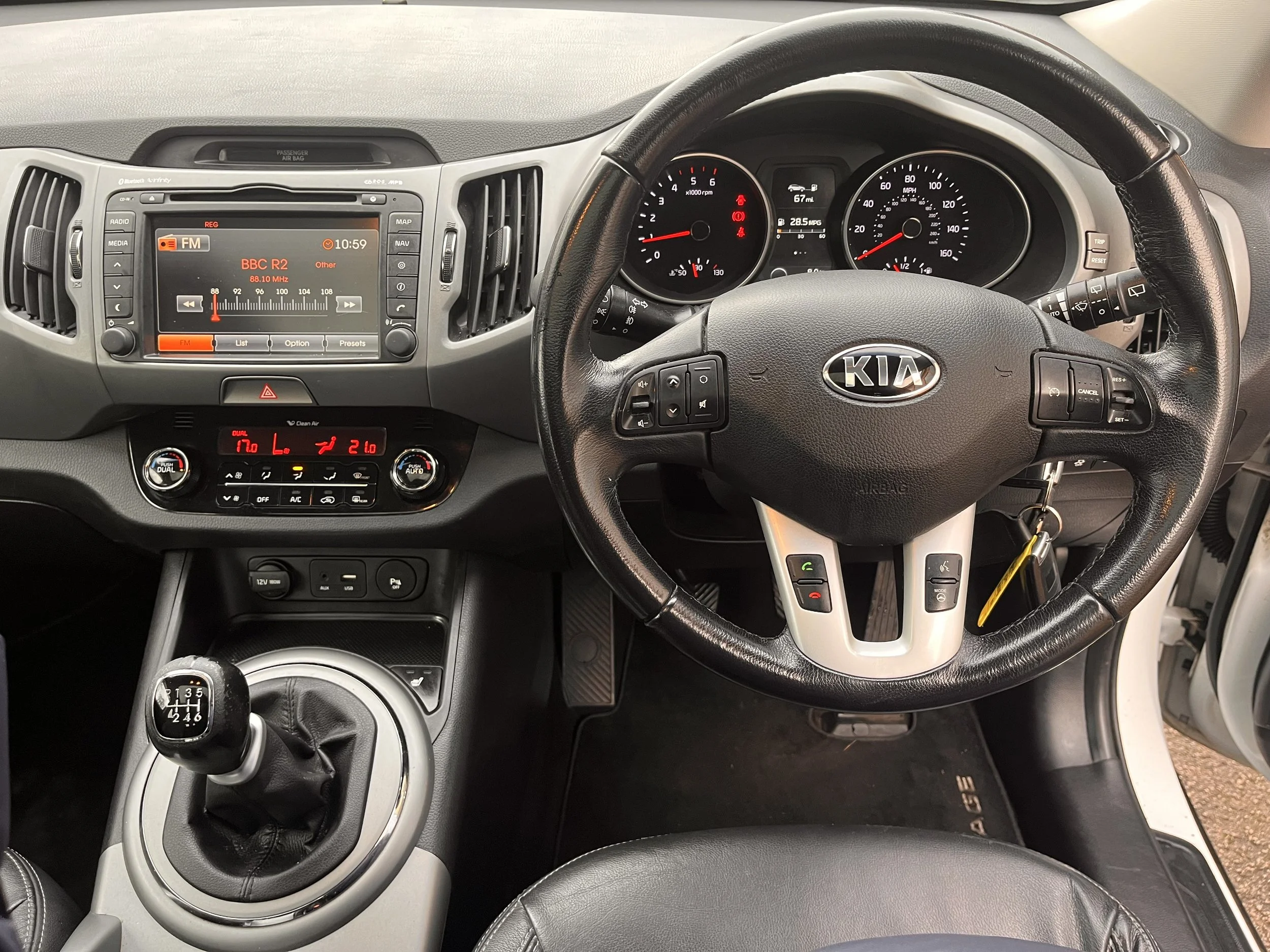The interior of a Kia vehicle dashboard showing a steering wheel, gear shift, and various controls and instruments, including a digital display tuned to BBC R2 on the radio.