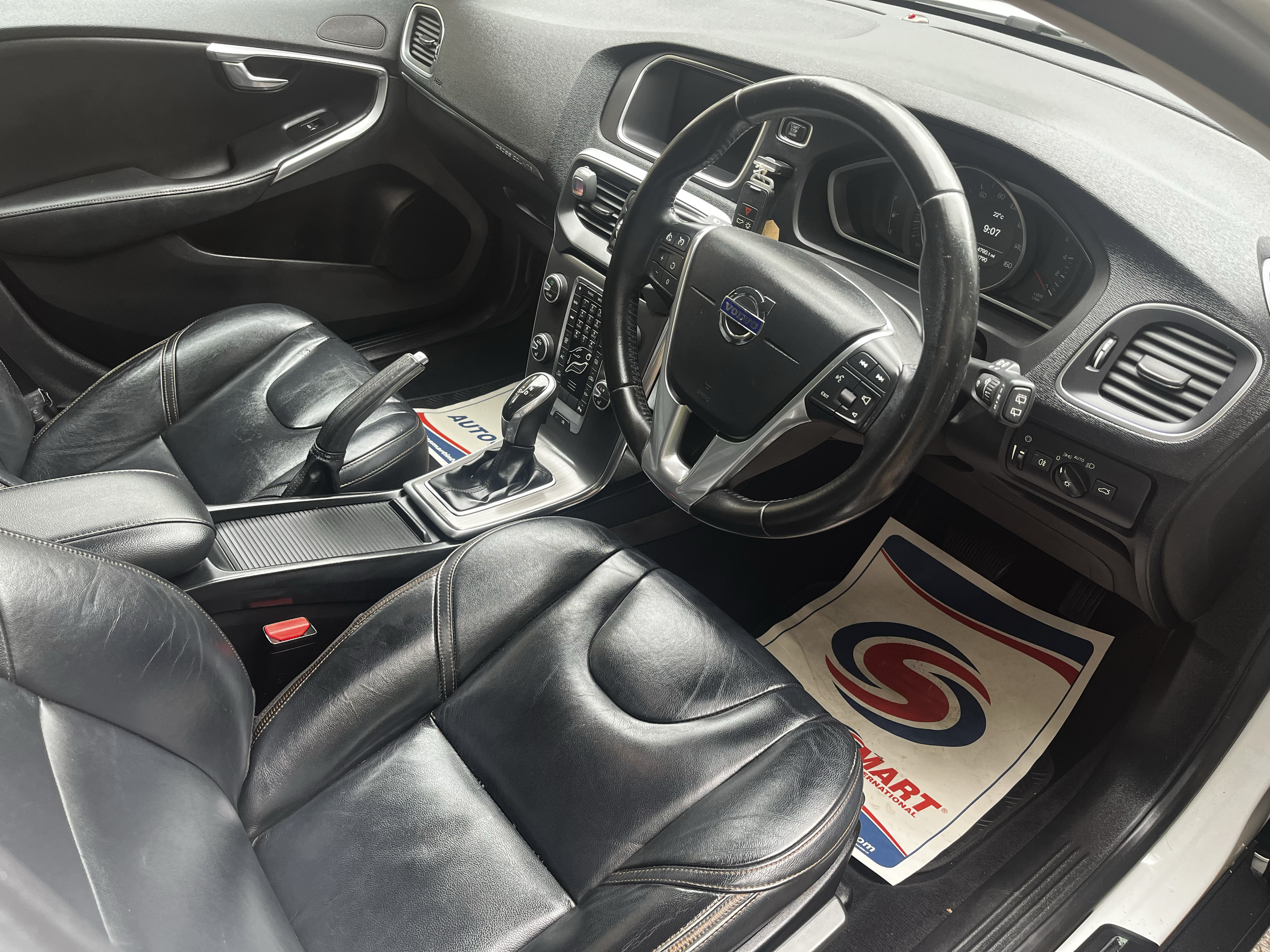 Interior of a black car showing the dashboard, steering wheel with Volvo logo, gear shifter, black leather seats, and floor mat with a red, white, and blue design.