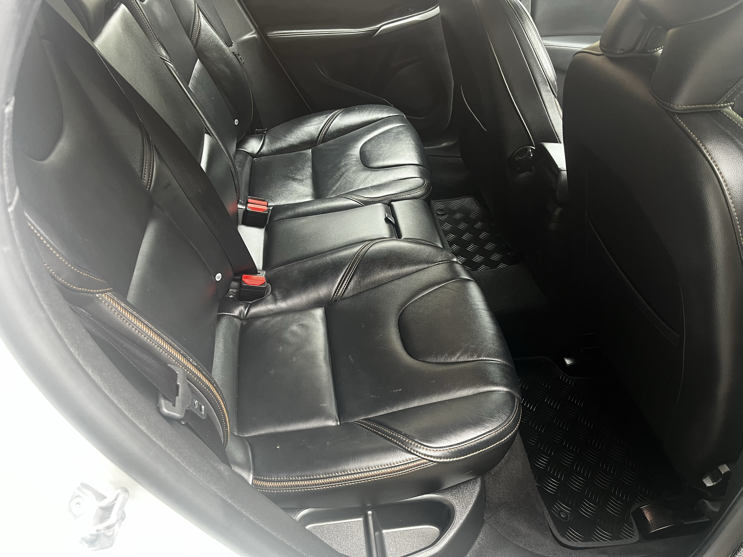 The backseat area of a car featuring black leather seats with contrast stitching, seatbelt buckles visible, and a black rubber floor mat with a textured pattern.