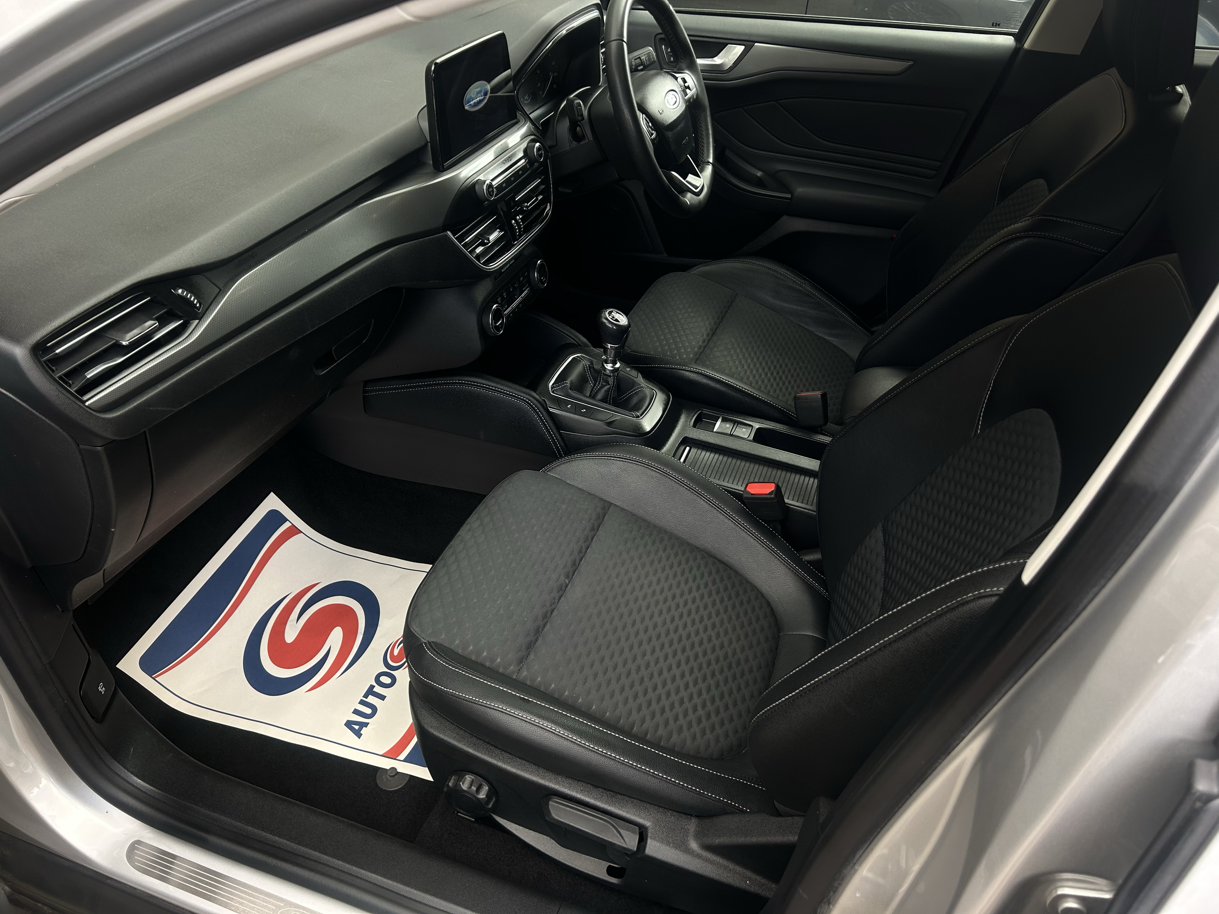 Interior of a modern car showing the driver's seat, steering wheel, dashboard, center console, and gear shift, with a paper floor mat on the driver's side.