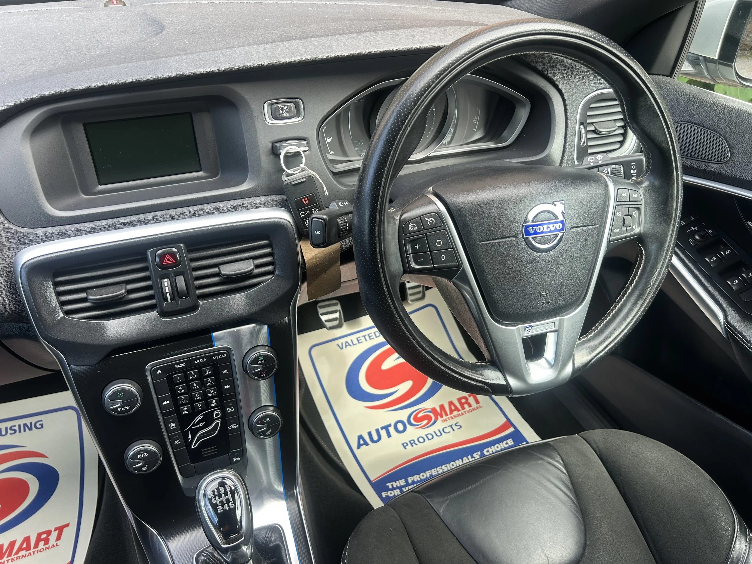 Inside view of a Volvo car's dashboard and steering wheel, showing controls, gear shift, and a sales promotional mat on the floor.
