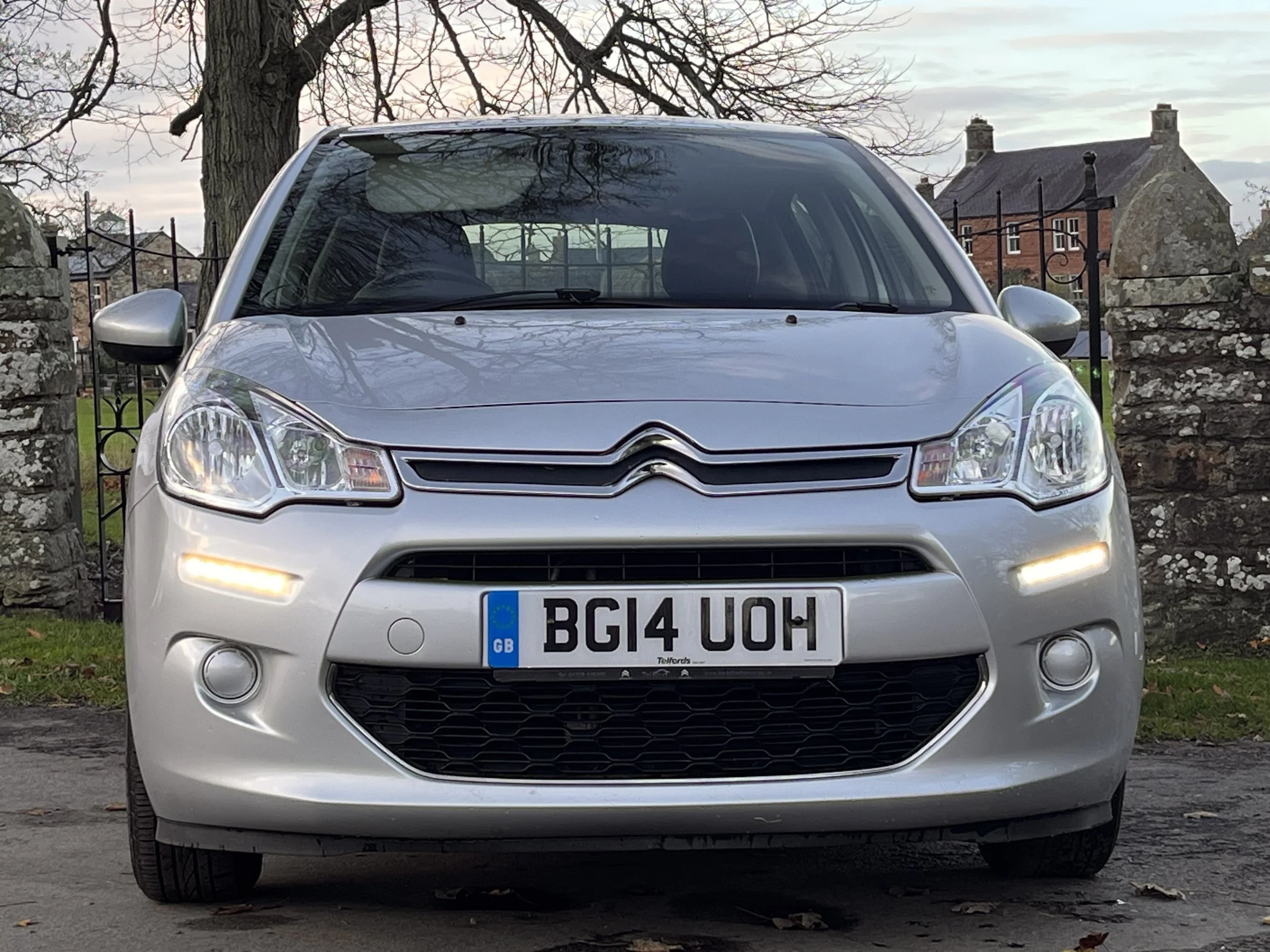 Front view of a silver Citroën car parked outdoors with a decorative metal fence, stone wall, and houses in the background.
