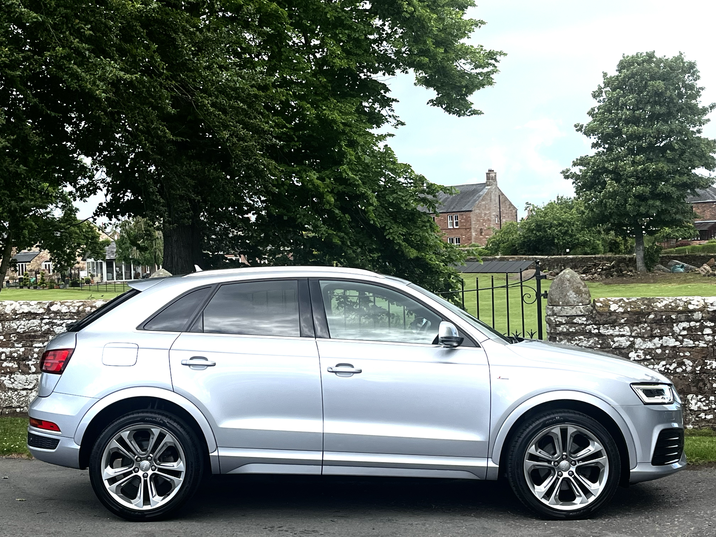 A silver SUV parked in front of a stone wall with trees and a historic red-brick building in the background.