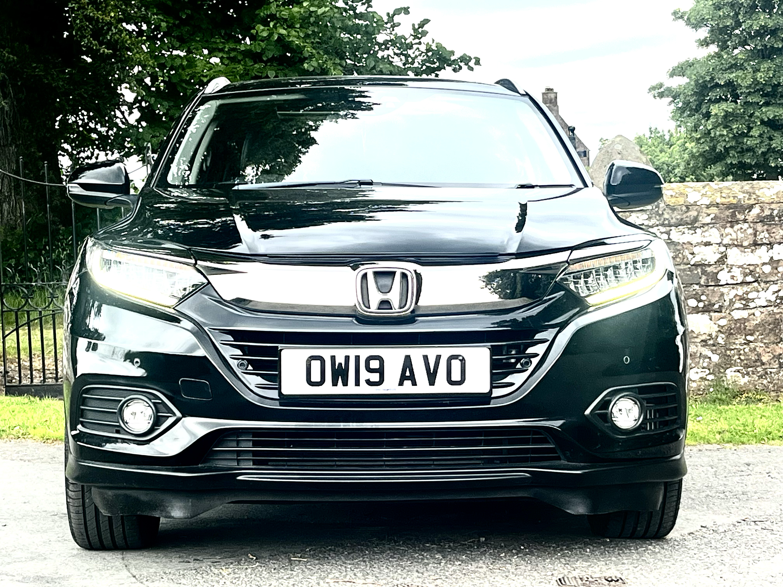 Front view of a black Honda SUV with a UK license plate, parked in a outdoor setting with trees and stone walls in the background.