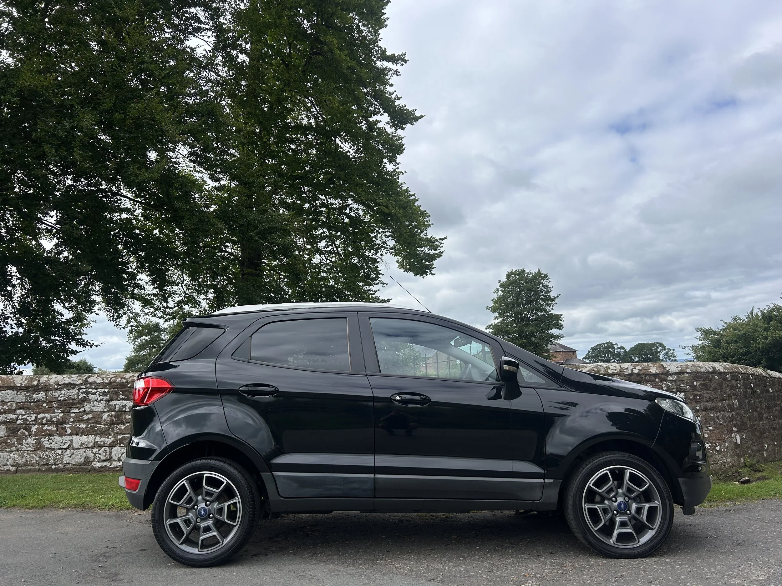 Black compact SUV parked on a road with a stone wall and green trees in the background under a cloudy sky.