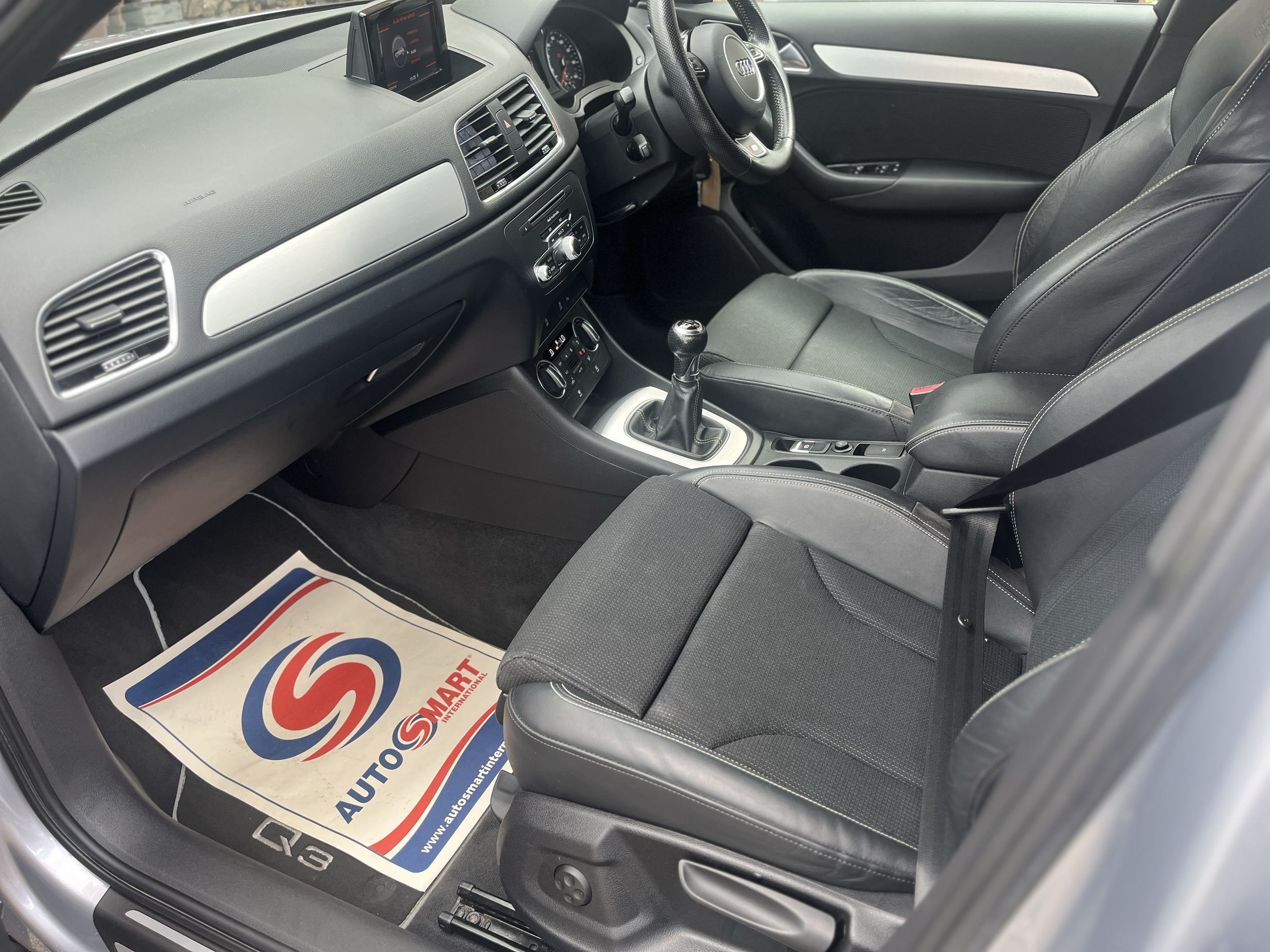 The interior of a car showing the driver's seat, steering wheel, dashboard with controls, and a gear shift. There is a paper floor mat with a logo on the floor.