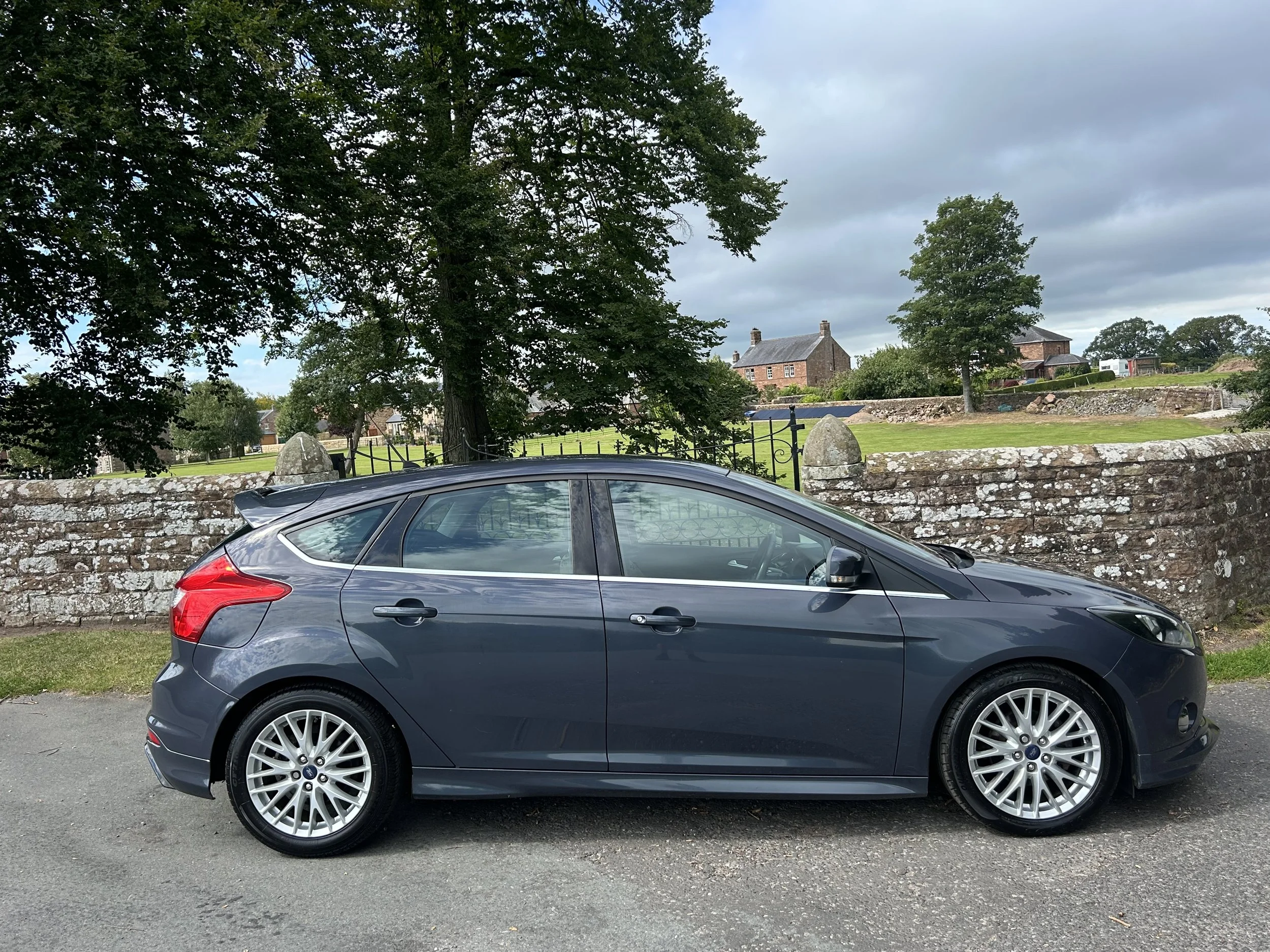 A dark gray hatchback car parked beside a stone wall with houses and trees in the background under a cloudy sky.