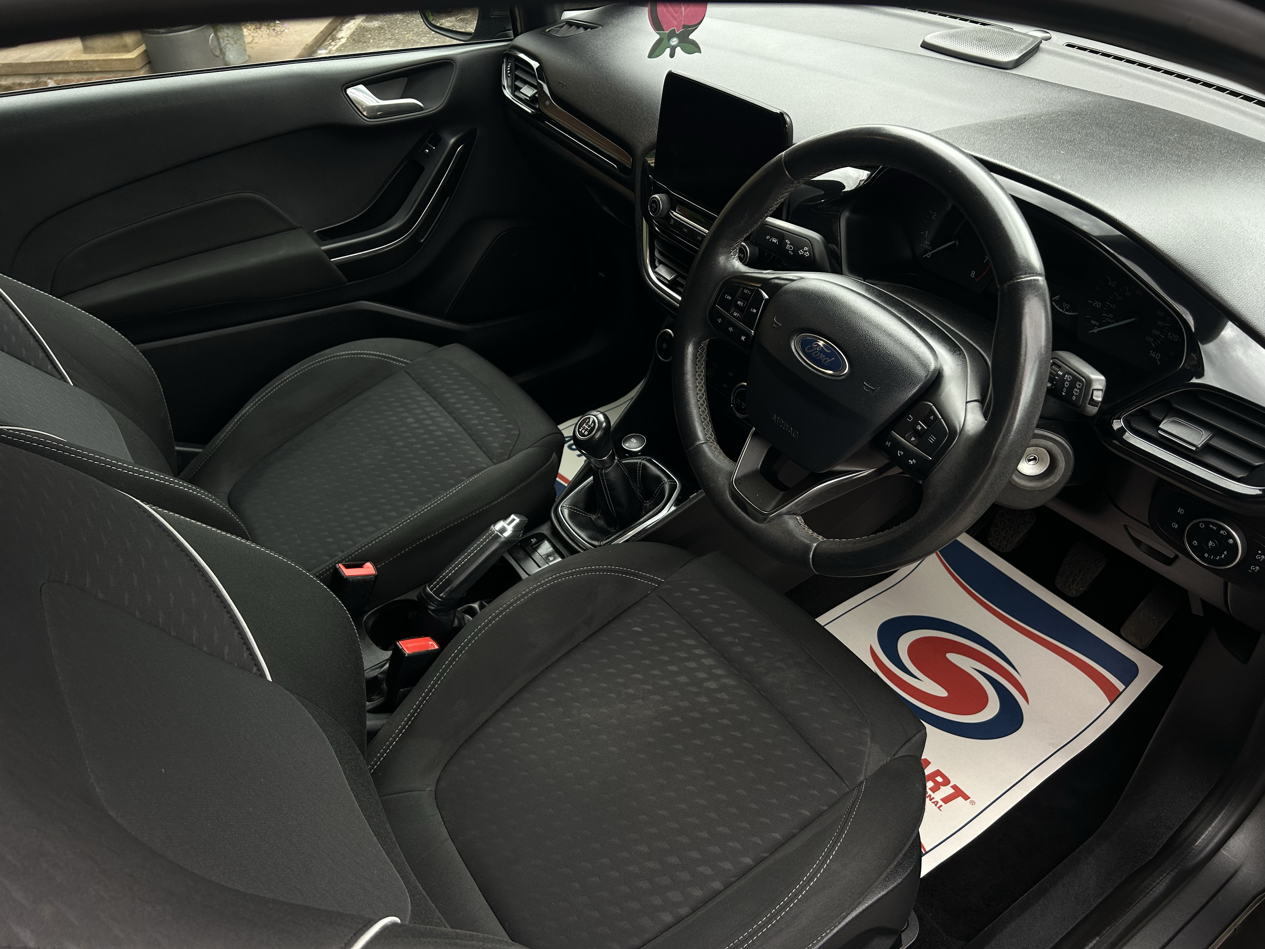 The interior of a black Ford vehicle showing the driver's side, a steering wheel, dashboard with display, and dark fabric seats. A logo and part of a paper mat with red and blue design are visible on the floor.