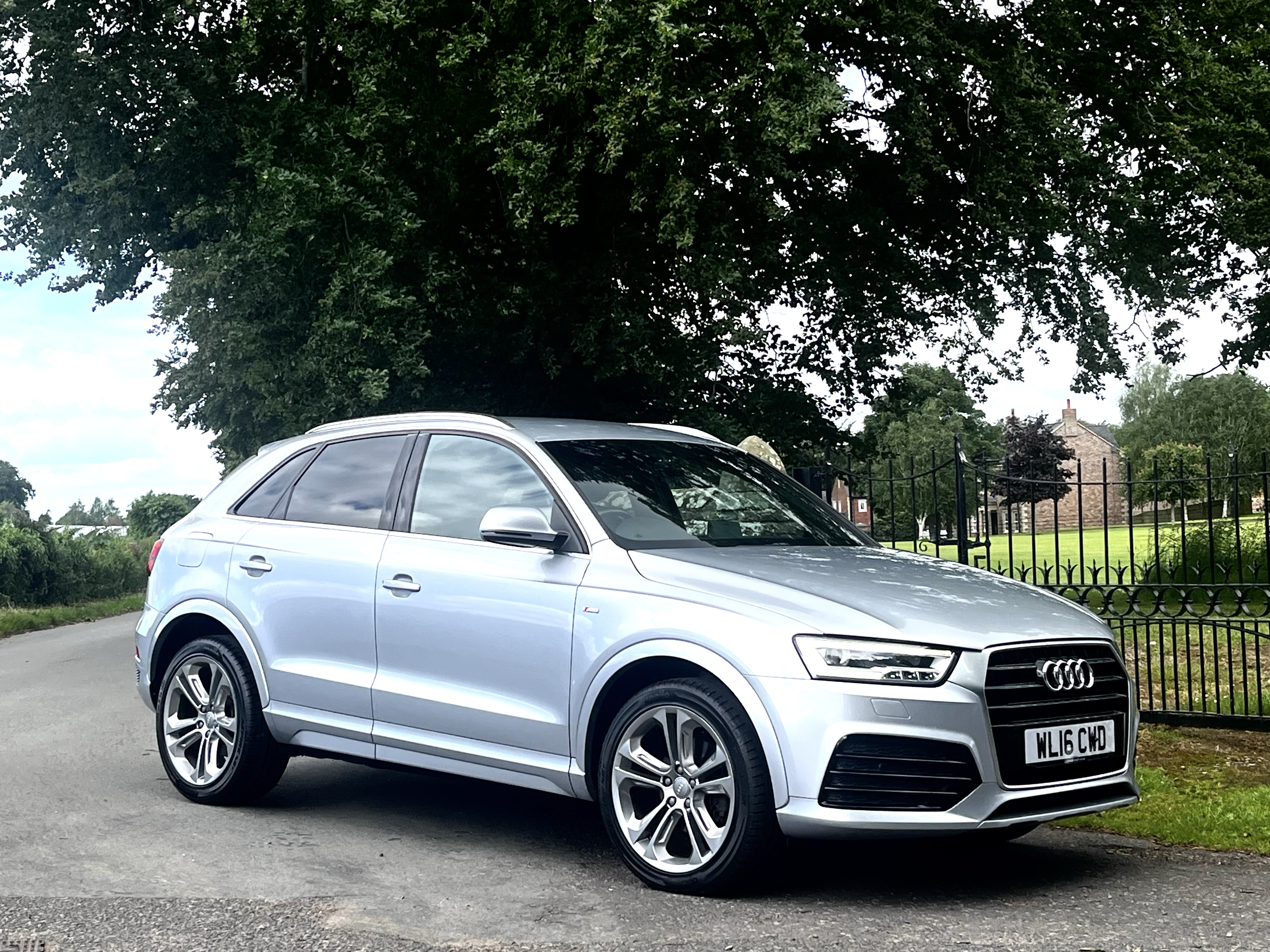 A silver Audi SUV parked on a street in front of a large tree with green leaves, a black metal fence, and houses in the background.