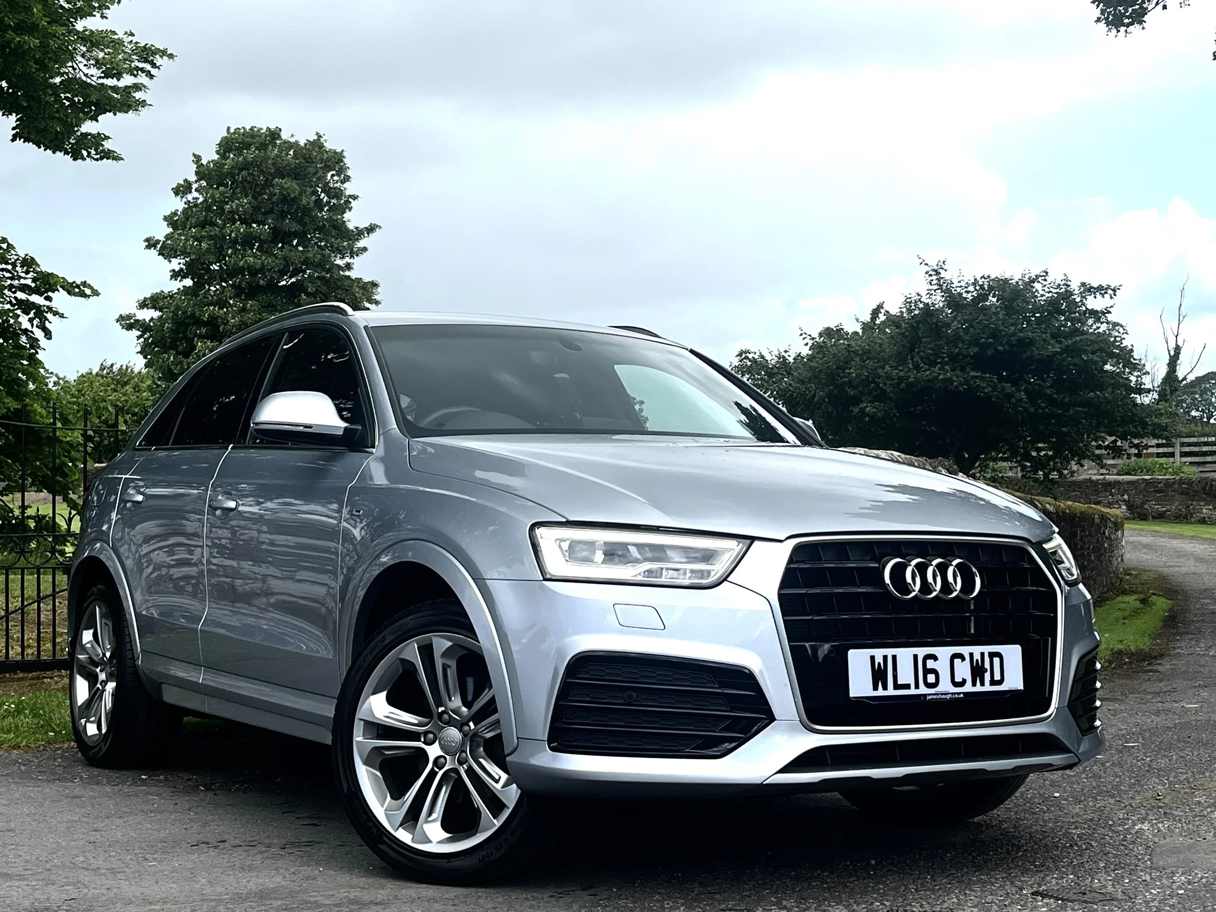 A silver Audi SUV parked outdoors on a paved surface, with trees, a fence, and cloudy sky in the background.