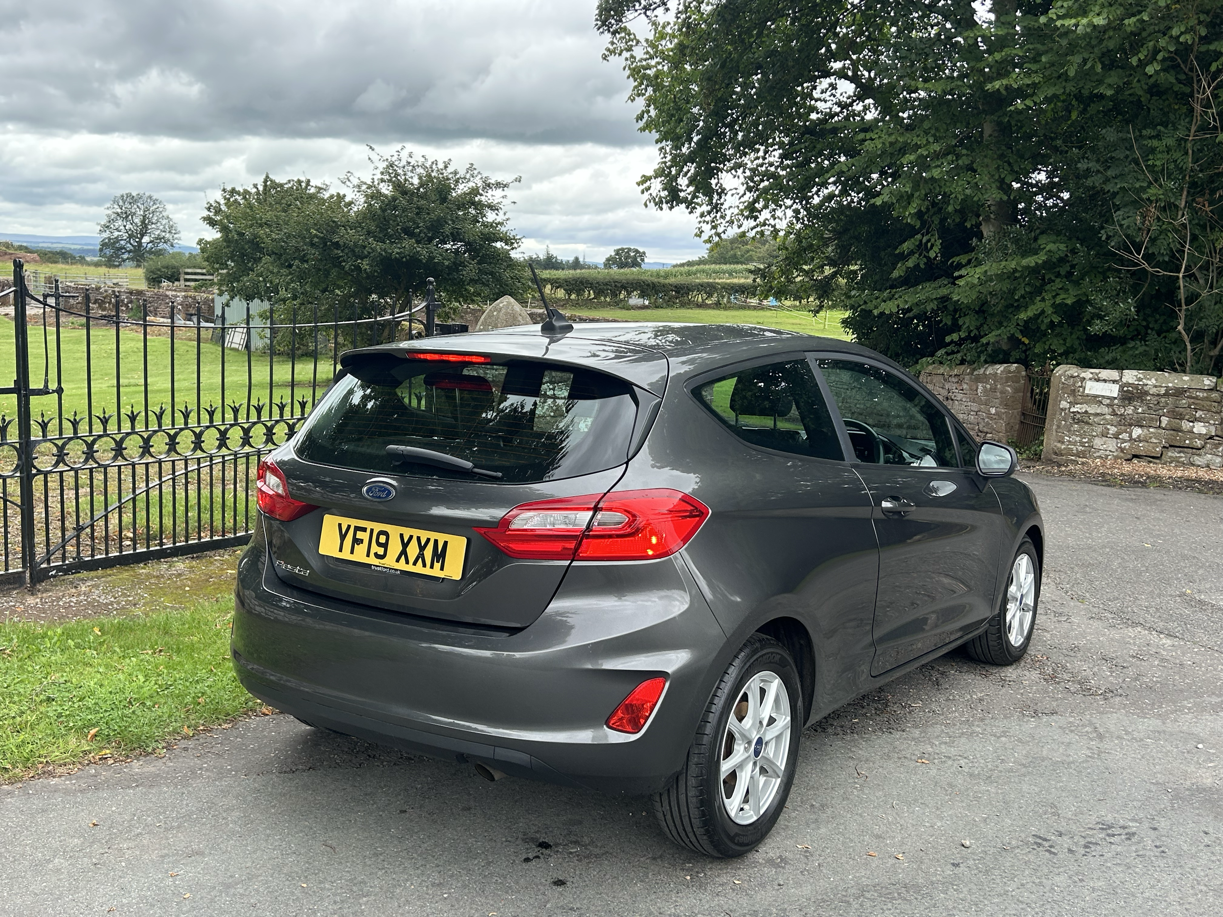 A grey Ford hatchback car parked on a paved surface. Behind the car is a black wrought iron fence, lush green grass, trees, and fields with overcast skies.
