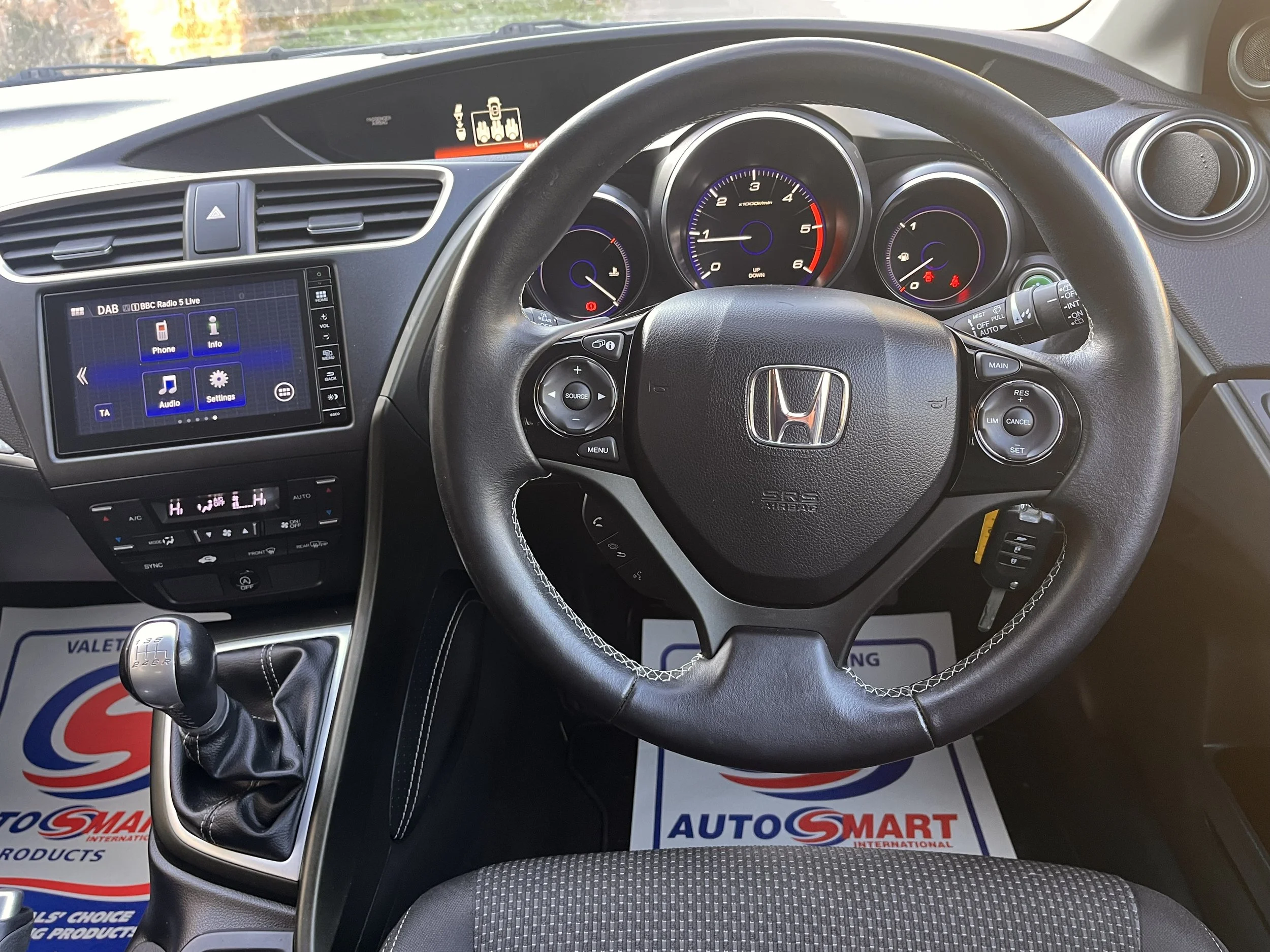 Inside the cockpit of a Honda vehicle showing the steering wheel, dashboard, gear shift, and infotainment system.