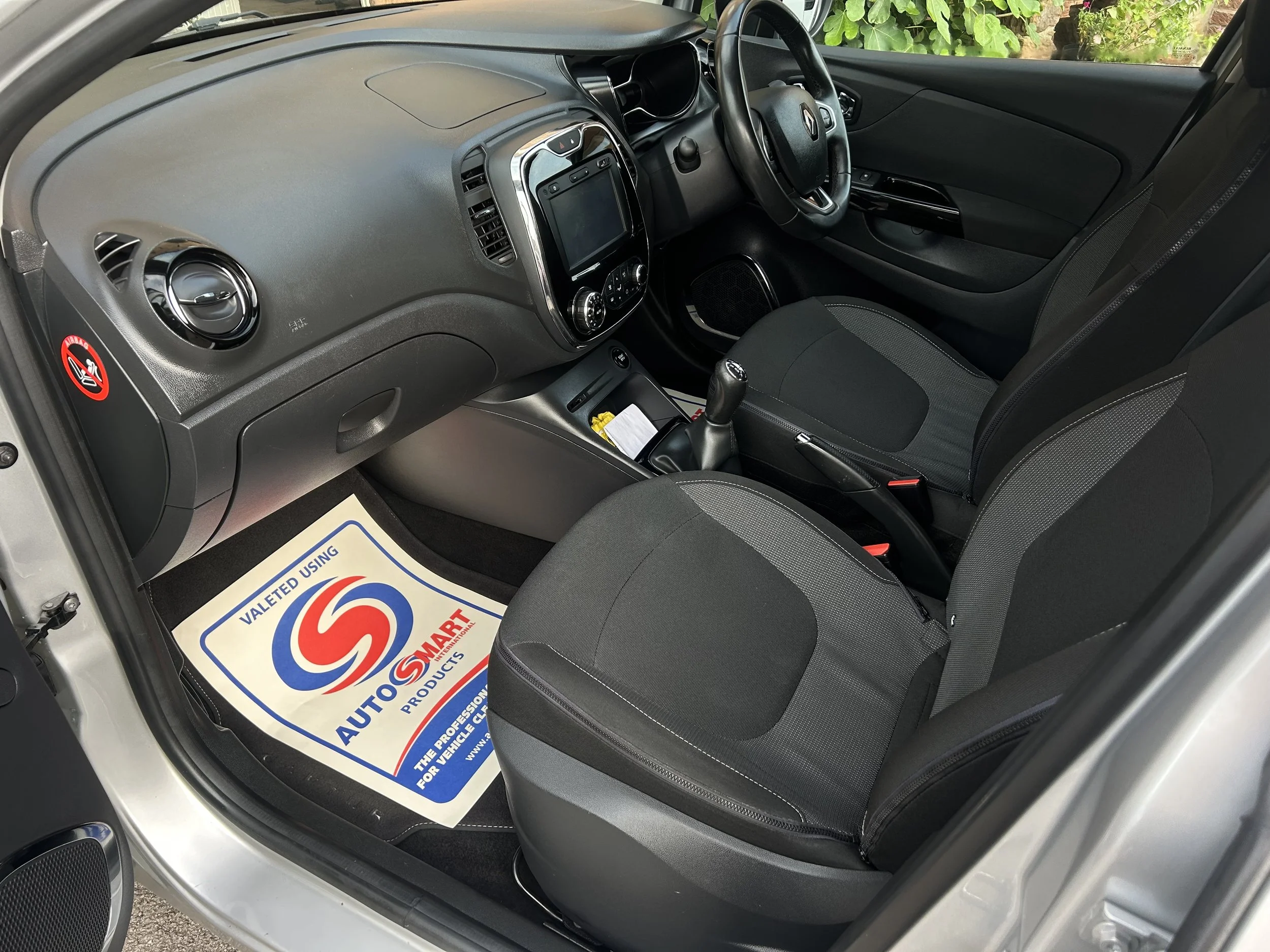 Interior of a small car showing the dashboard, steering wheel, gear shift, and front seats. The dashboard includes a display screen and air vents, with a no smoking sticker on the side. The floor has a paper mat with the AutoSmart logo.