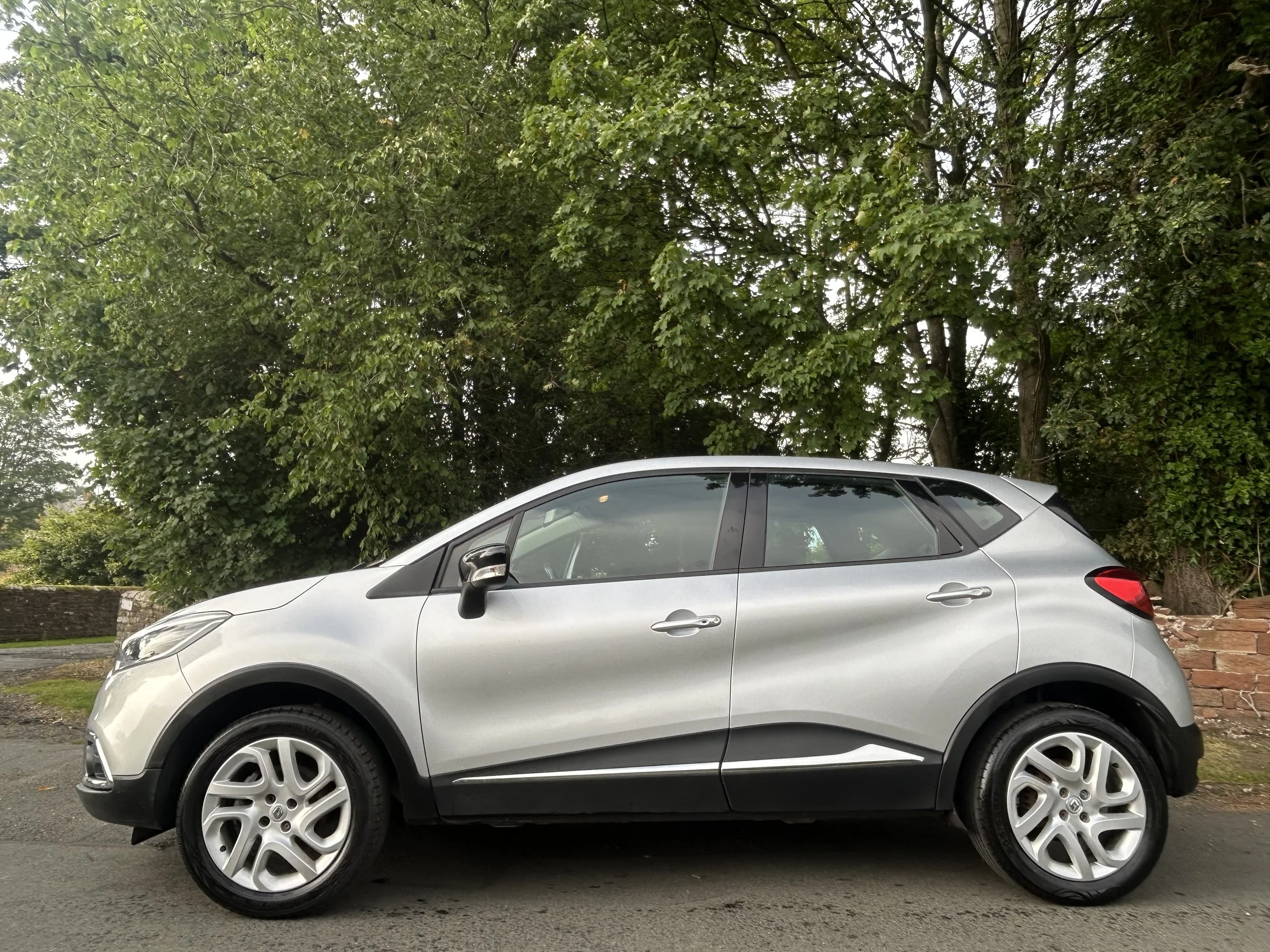 Side view of a silver compact crossover SUV parked on the street with green trees and a brick wall in the background.