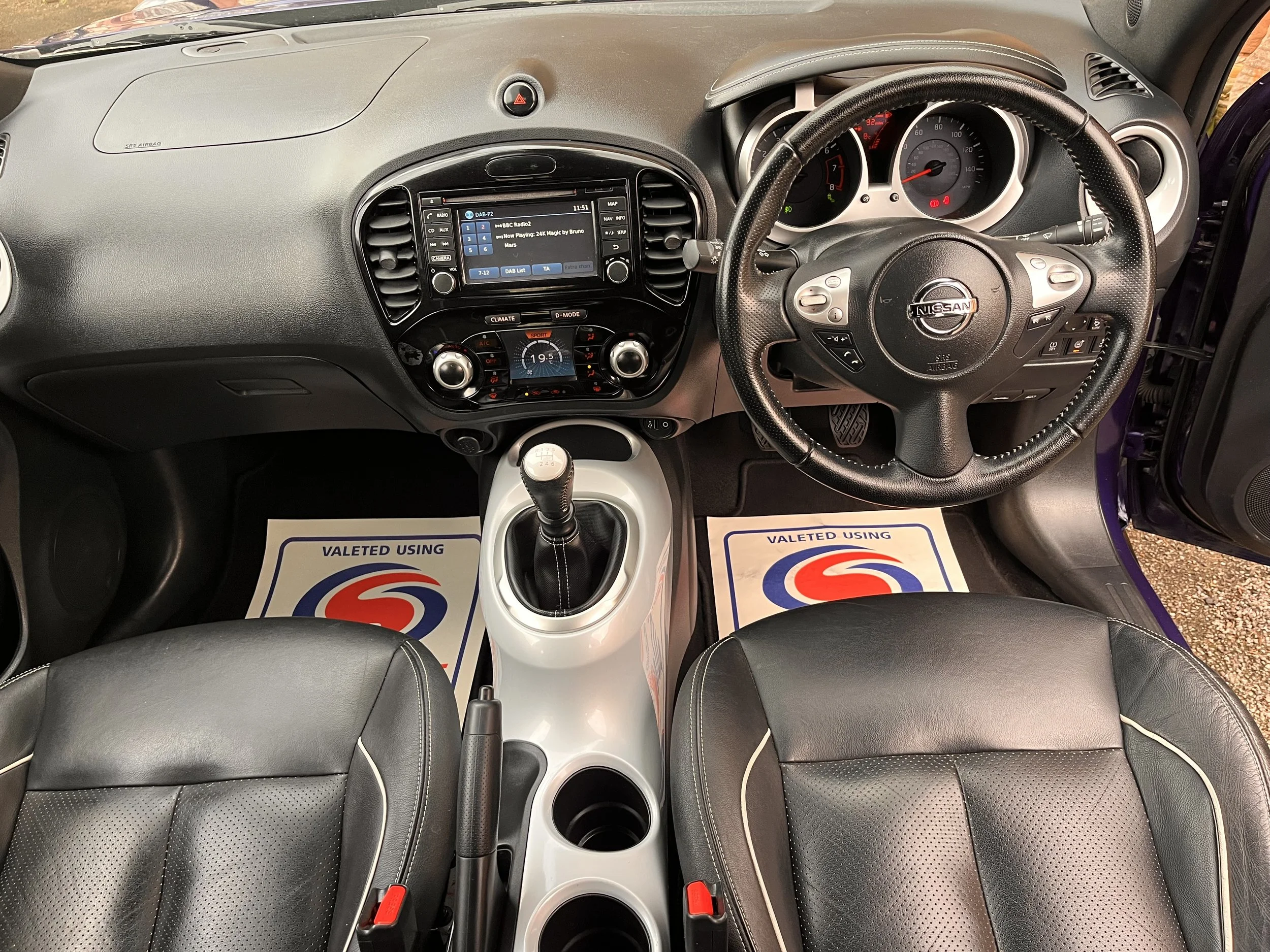 Front interior of a Nissan car showing the dashboard, steering wheel, gear shift, and two front seats with floor mats labeled 'Valeted Using'.