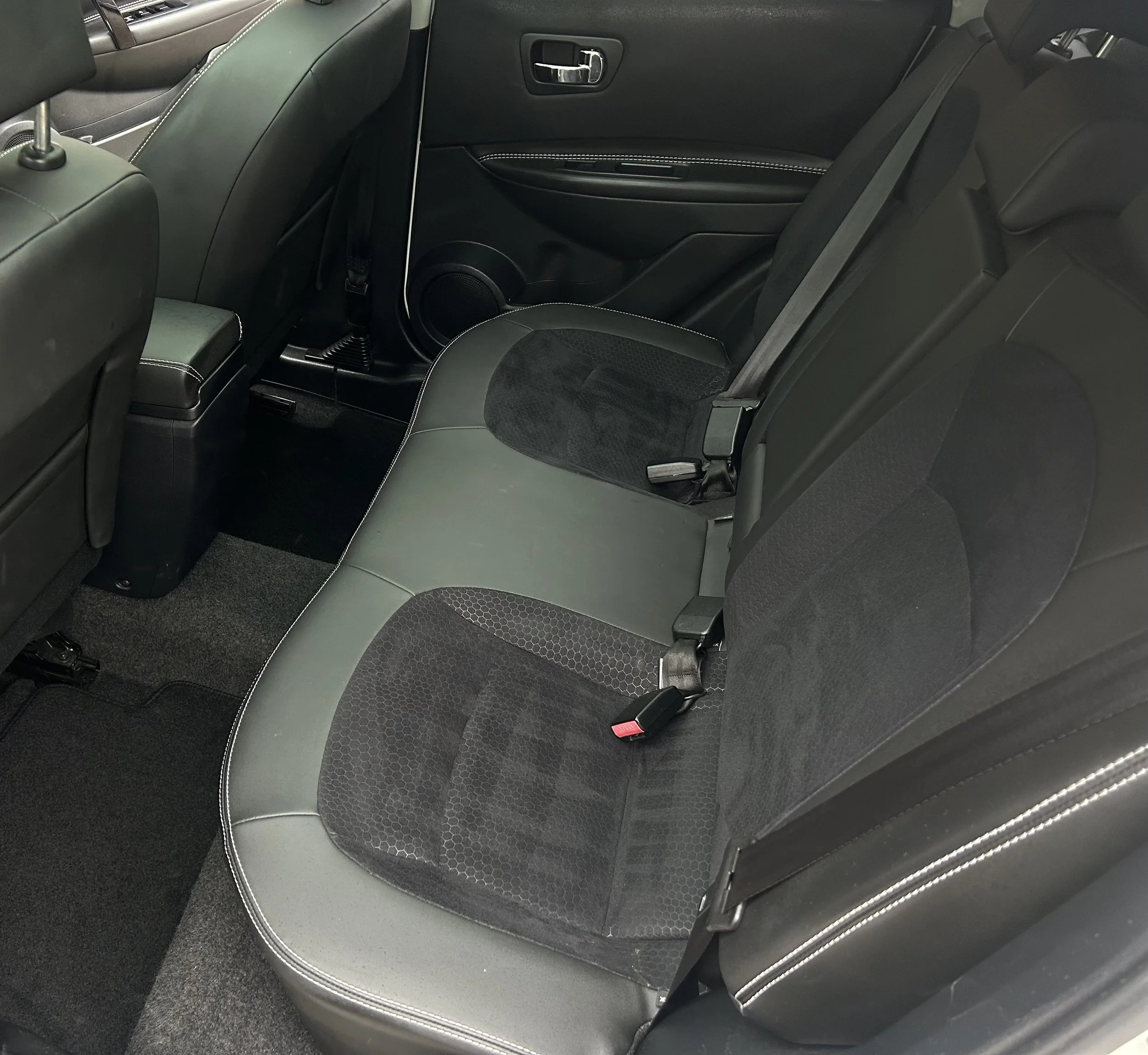 The backseat of a car featuring a black and gray cloth seat with seat belts and a door with black interior paneling.