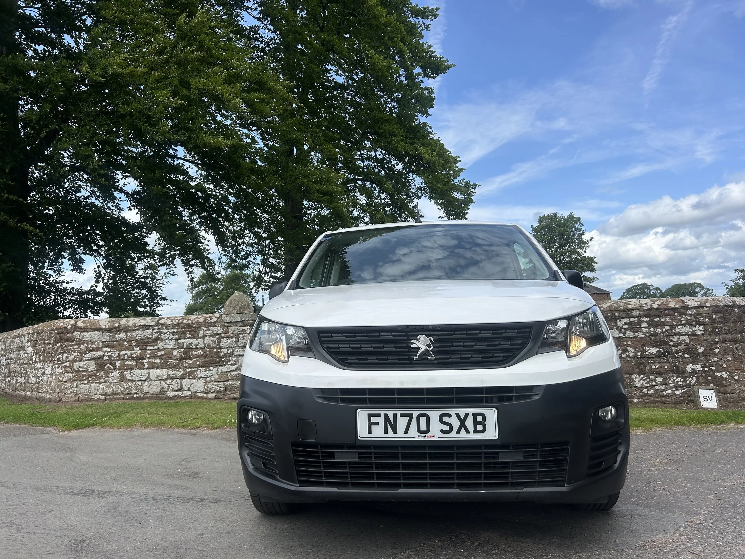 Front view of a white Peugeot car parked on a paved road, with a stone wall, green trees, and a partly cloudy sky in the background.