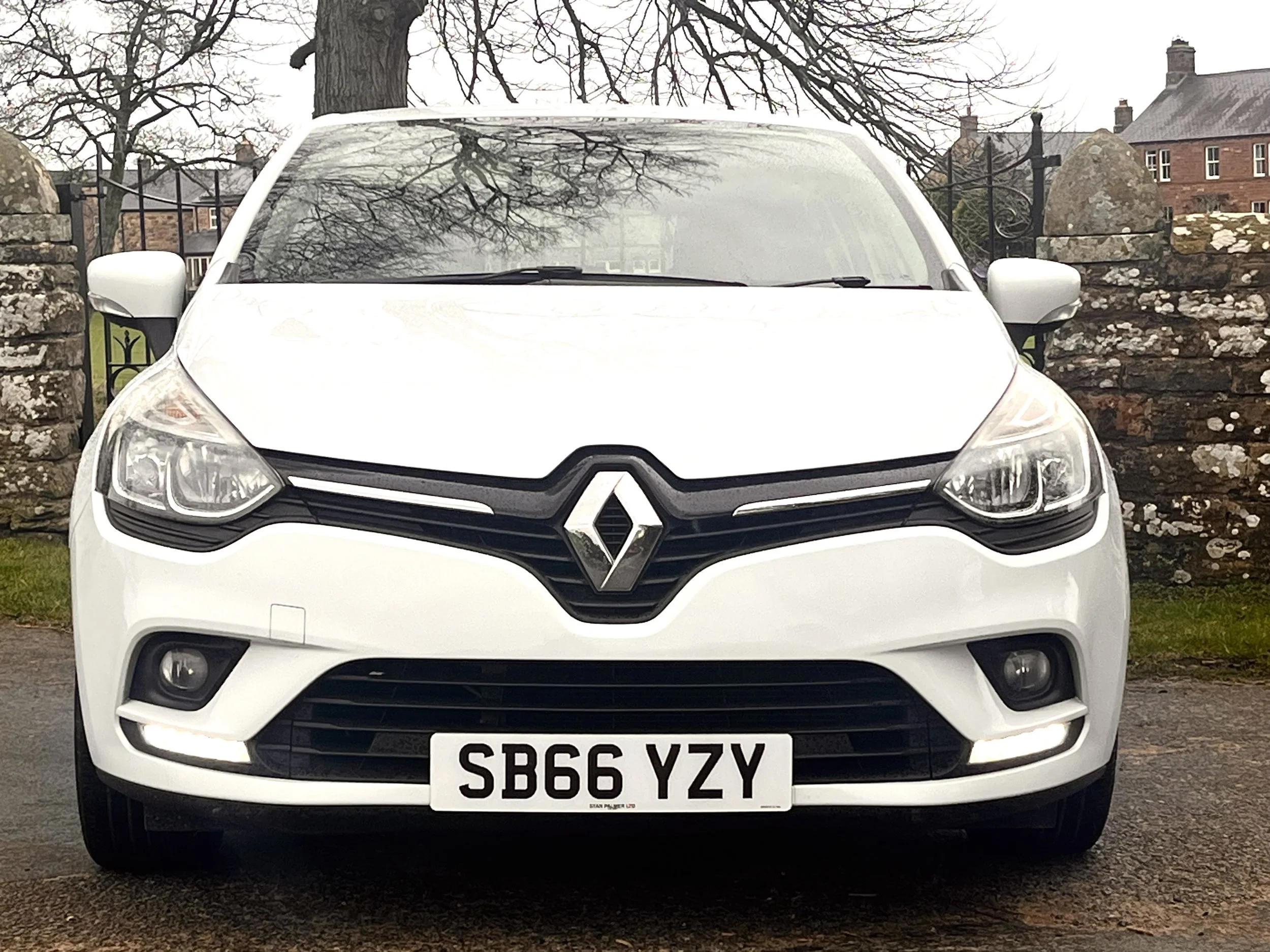 Front view of a white Renault car with a UK license plate, parked on a wet surface in front of a stone wall and trees, with cloudy sky and buildings in background.