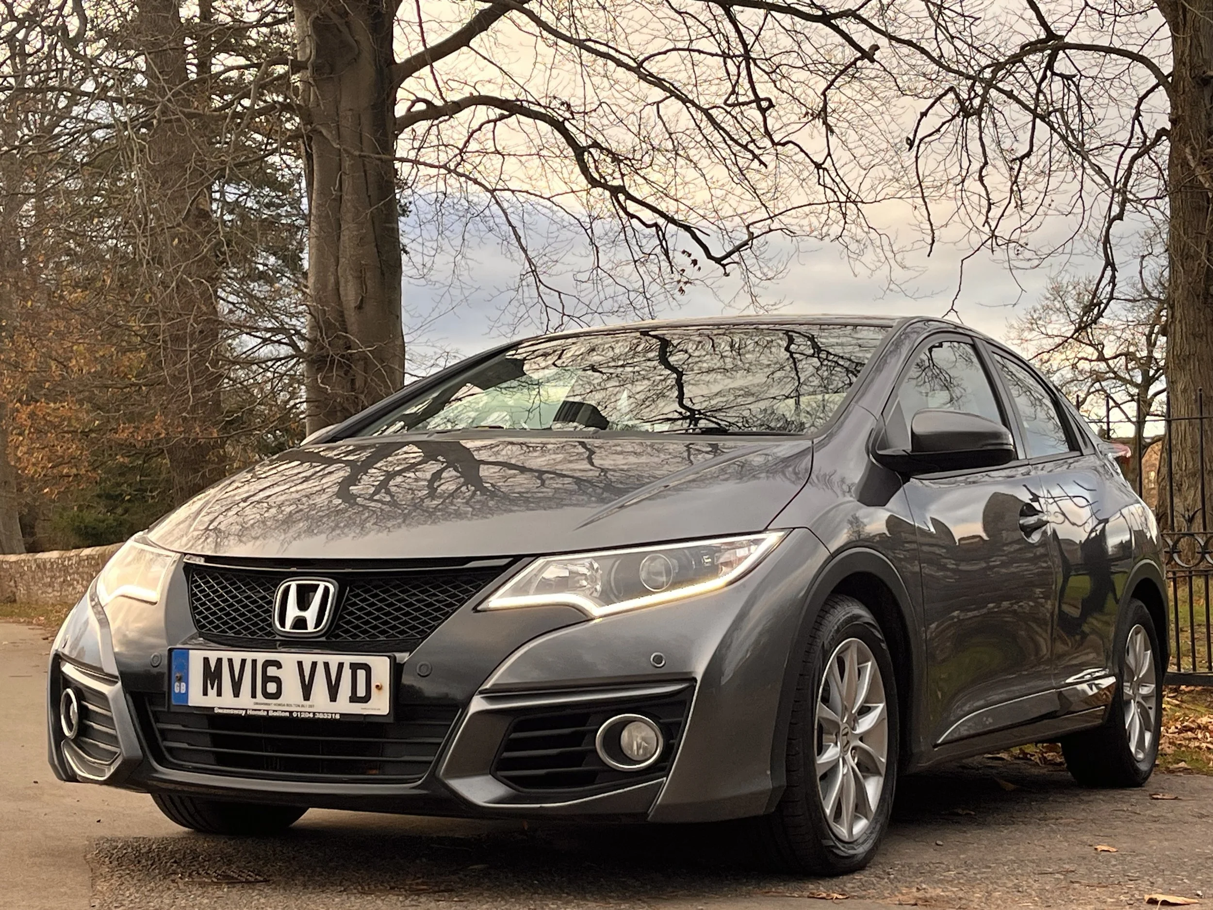 A gray Honda Civic car parked on a street with trees and a fence in the background during autumn.