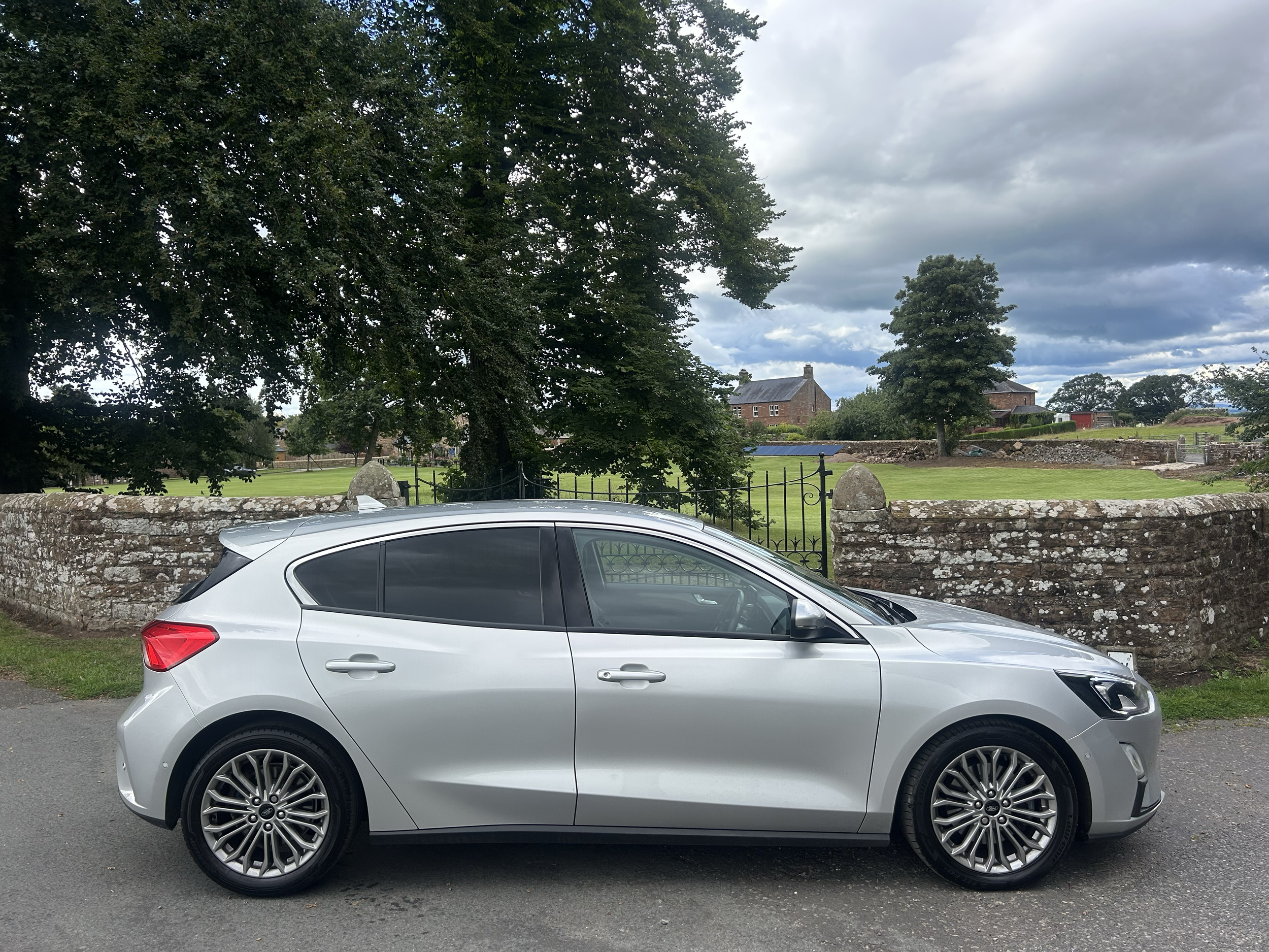 A silver car parked on a street in front of a stone wall with greenery and trees, and a stone house in the background under a cloudy sky.