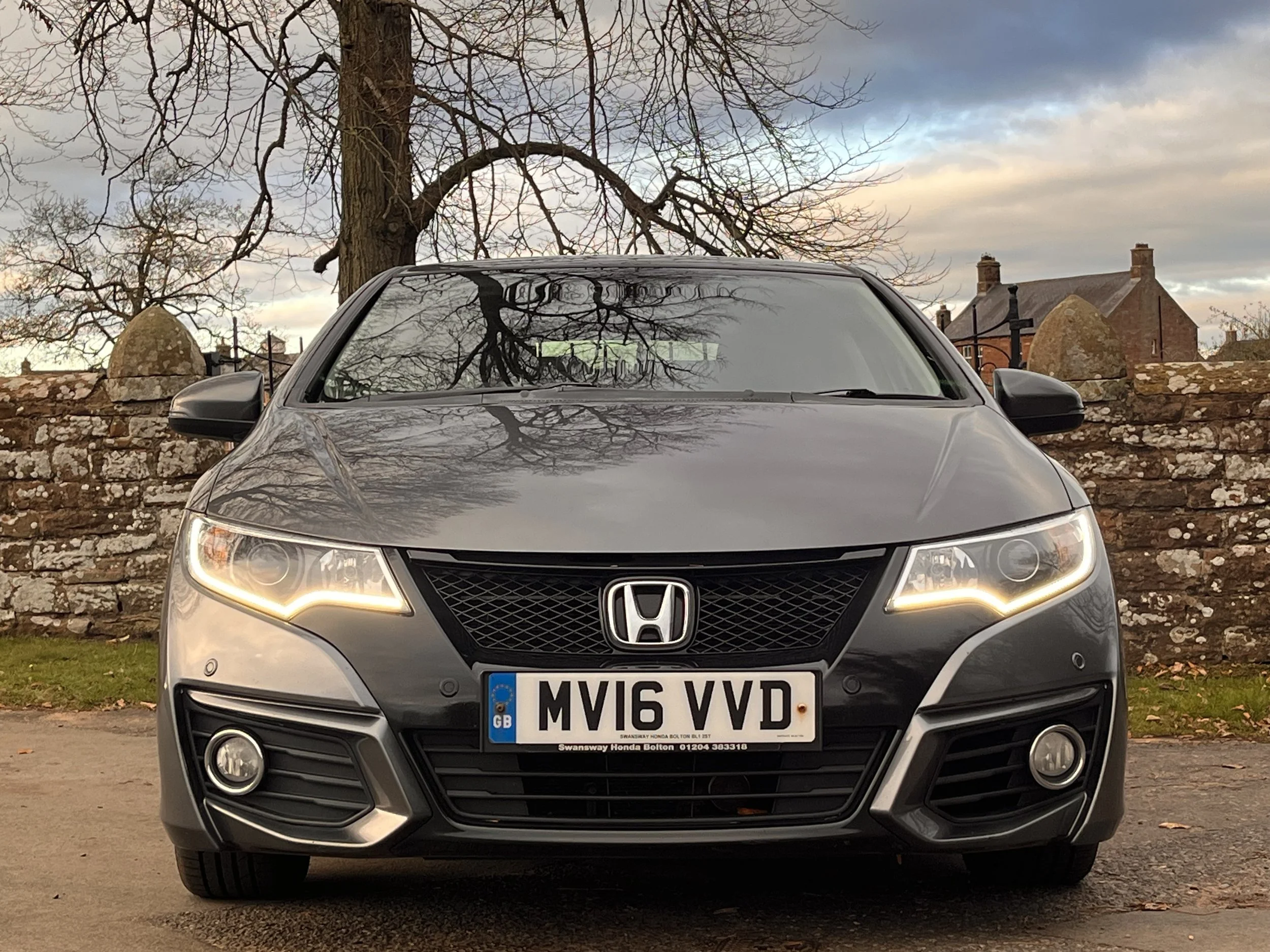 Front view of a gray Honda car with a UK license plate, parked outdoors with a stone wall, leafless tree, and houses in the background during cloudy weather.