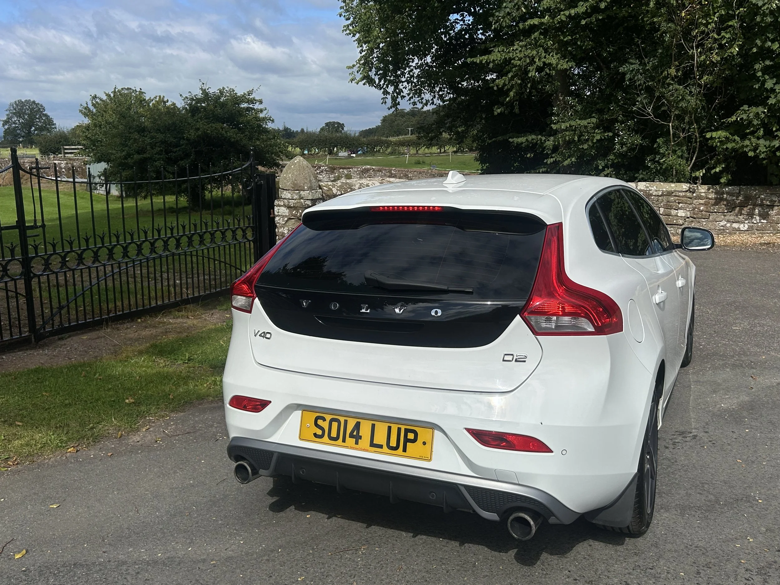 White Volvo V40 hatchback car parked on a paved driveway near a black metal gate and a stone wall, with green grass, trees, and a cloudy sky in the background.