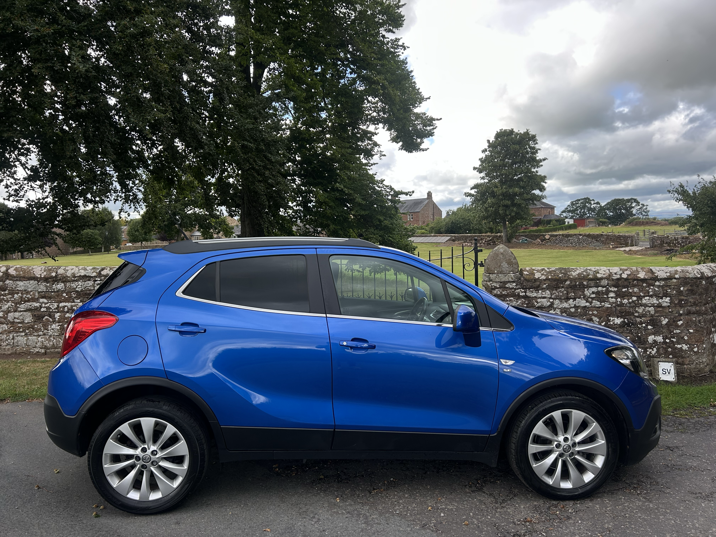 A bright blue compact SUV parked on a street with a stone wall and a grassy park area with trees and houses in the background, under a mostly cloudy sky.