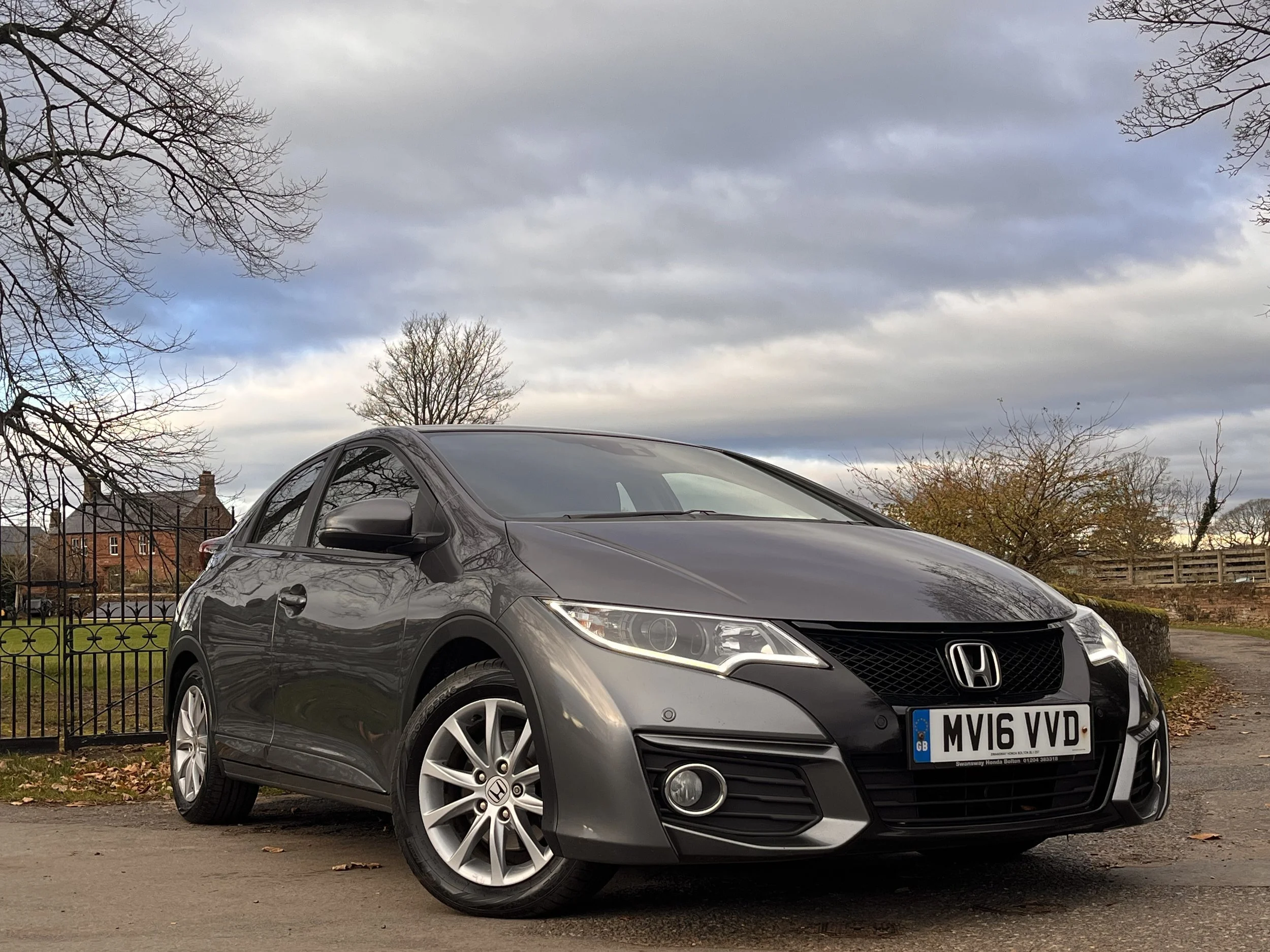 A gray Honda Civic parked on a road with trees and houses in the background under a cloudy sky.