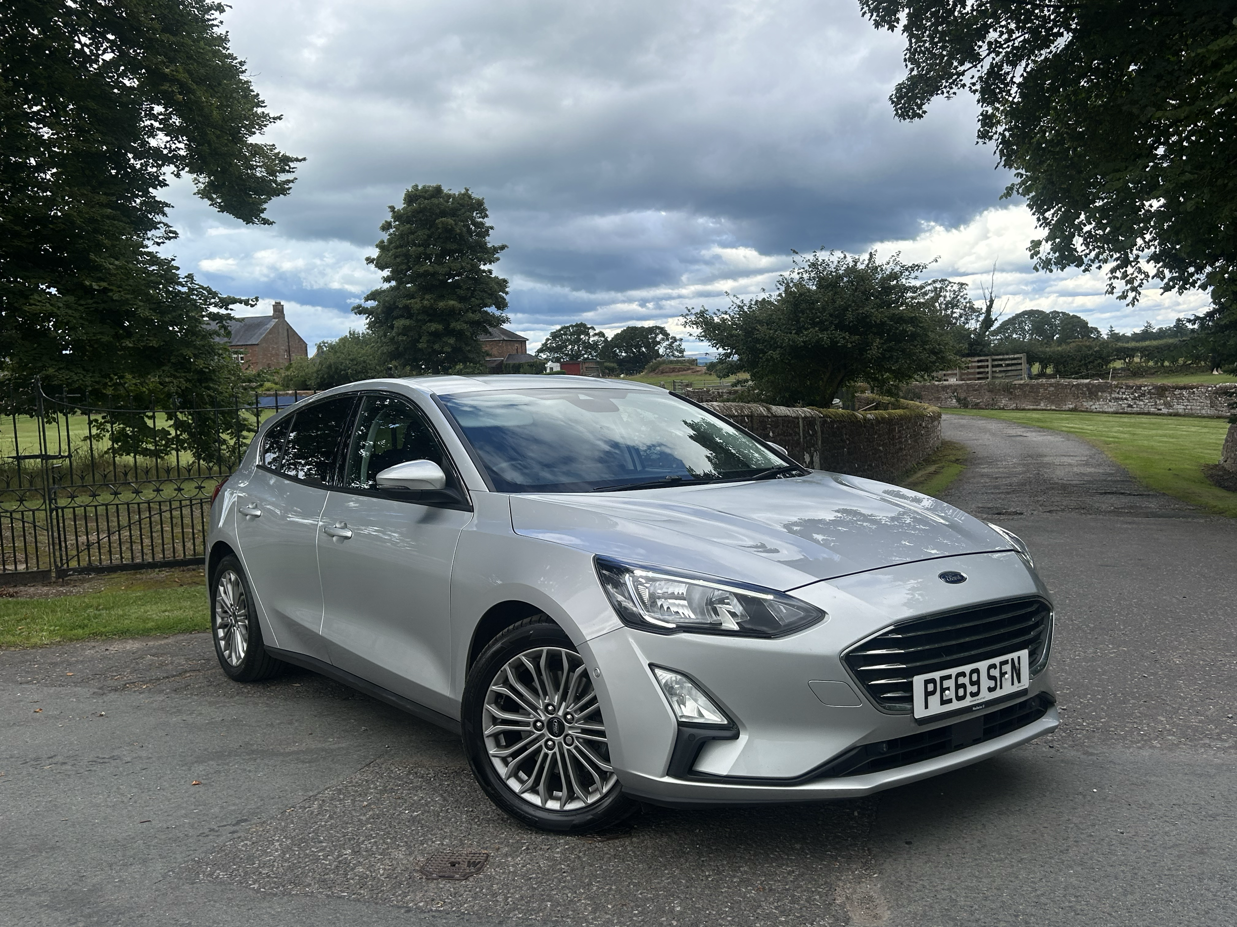 A silver Hyundai i40 parked on a driveway with a country house and trees in the background on a cloudy day.